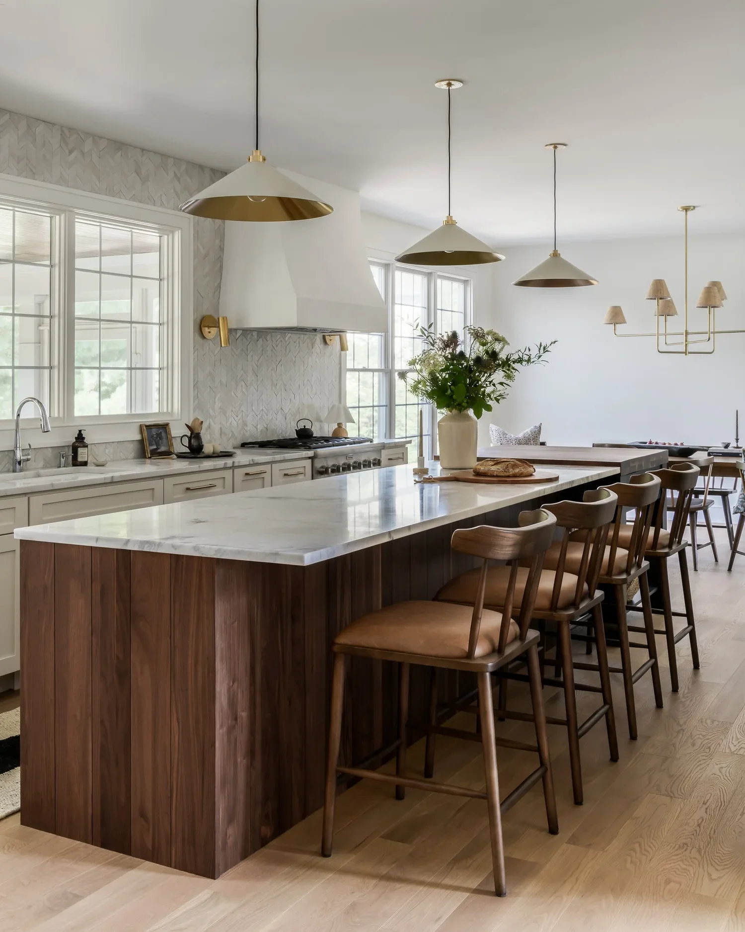 Modern kitchen with white cabinetry, marble countertops, a large island with wooden base, and barstools. Pendant lights hang above the island. There are large windows, a tasteful arrangement of kitchenware, and a vase with flowers on the counter.