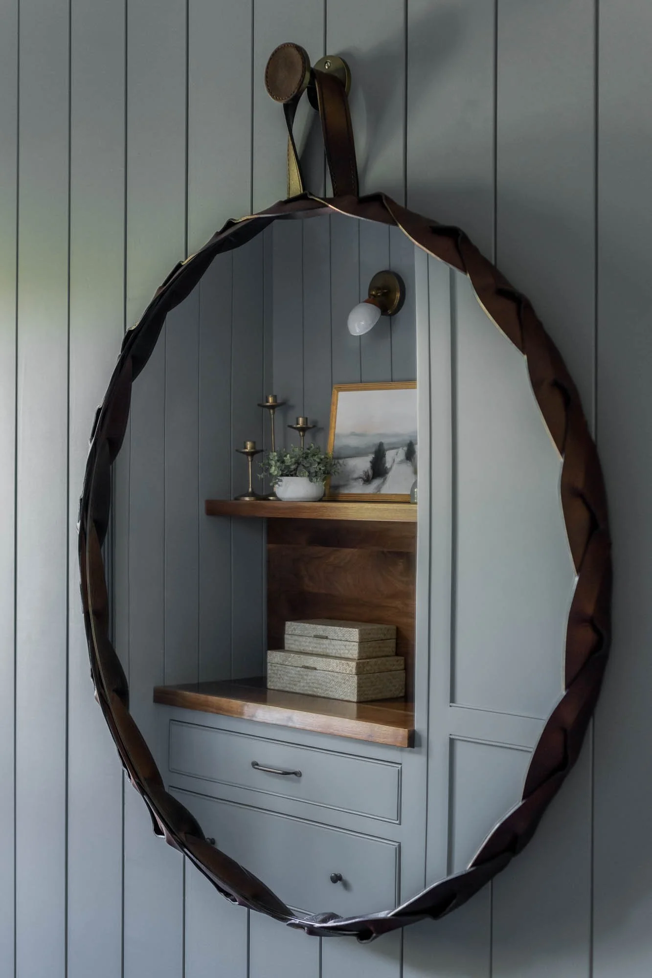 A mirror with a dark wood frame hanging on a gray paneled wall, reflecting a shelf with decor including candlesticks, a framed picture, a small plant, and stacked boxes.