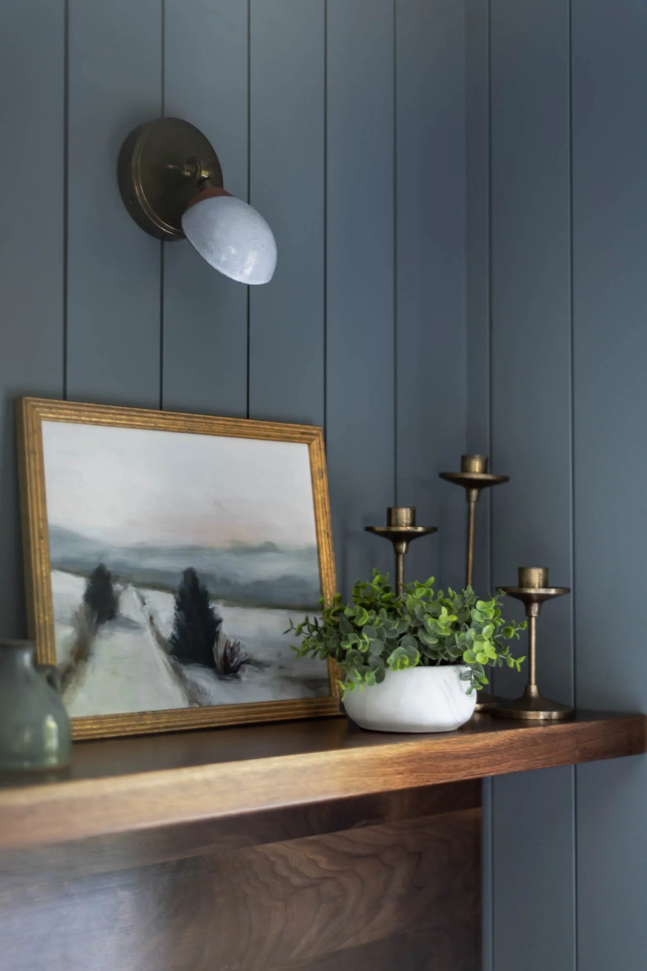 A blue wooden wall with a brass wall-mounted light fixture, a framed landscape painting, a white ceramic planter with green leafy plant, and a brass candle holder on a wooden surface.