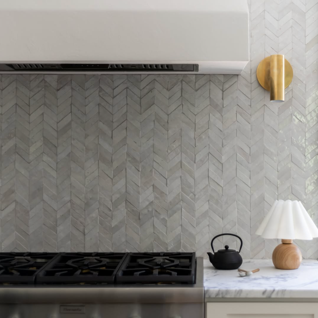 Kitchen with gray tile backsplash, black teapot, white lamp with wooden base on marble countertop, brass wall light.