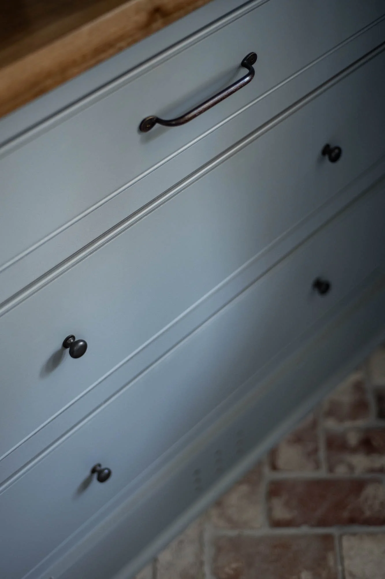 Close-up of a light gray wooden cabinet with black handles, situated on a brick floor.