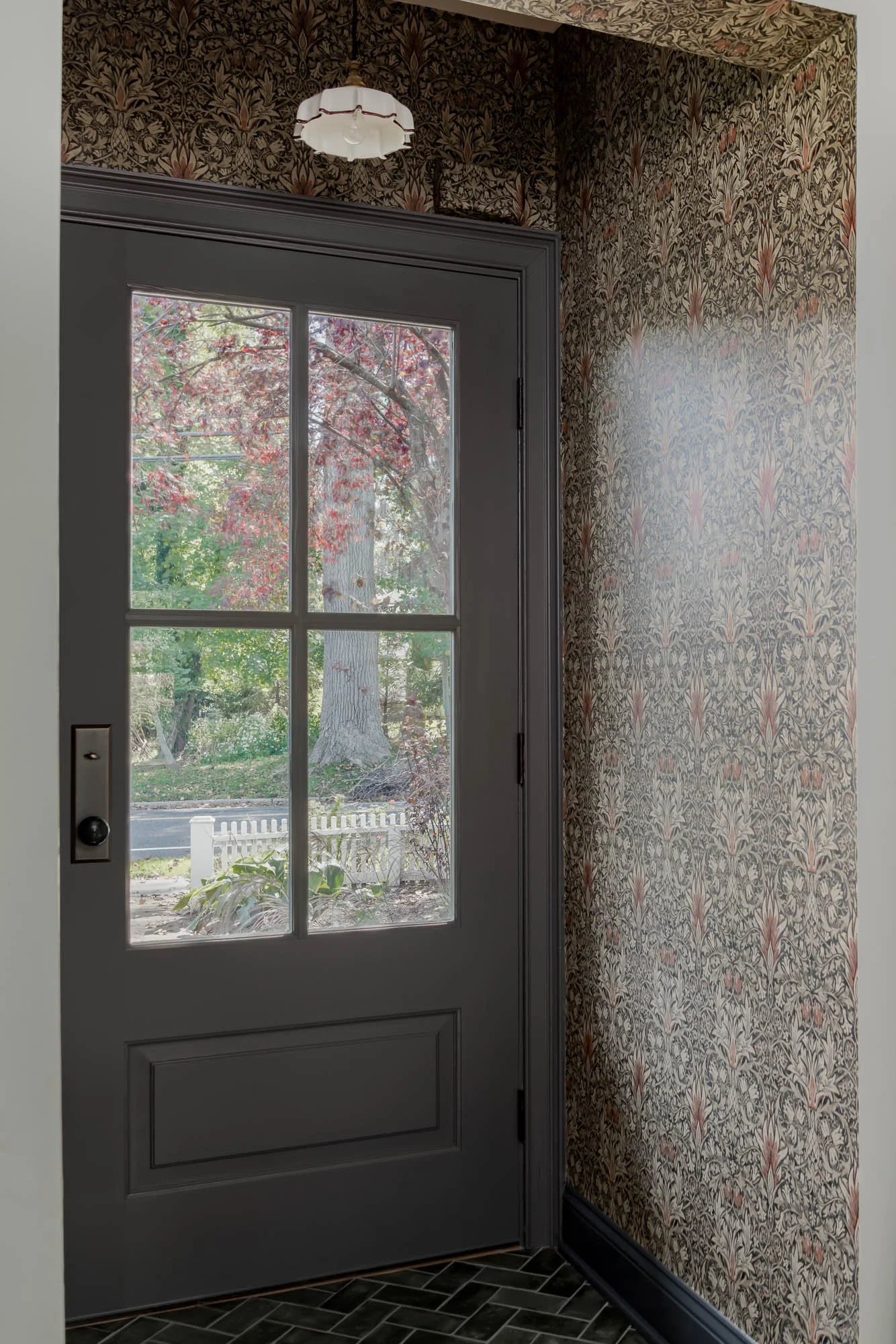 A door with glass panes and a floral-patterned wallpaper next to it. Outside, trees with red and green leaves are visible.