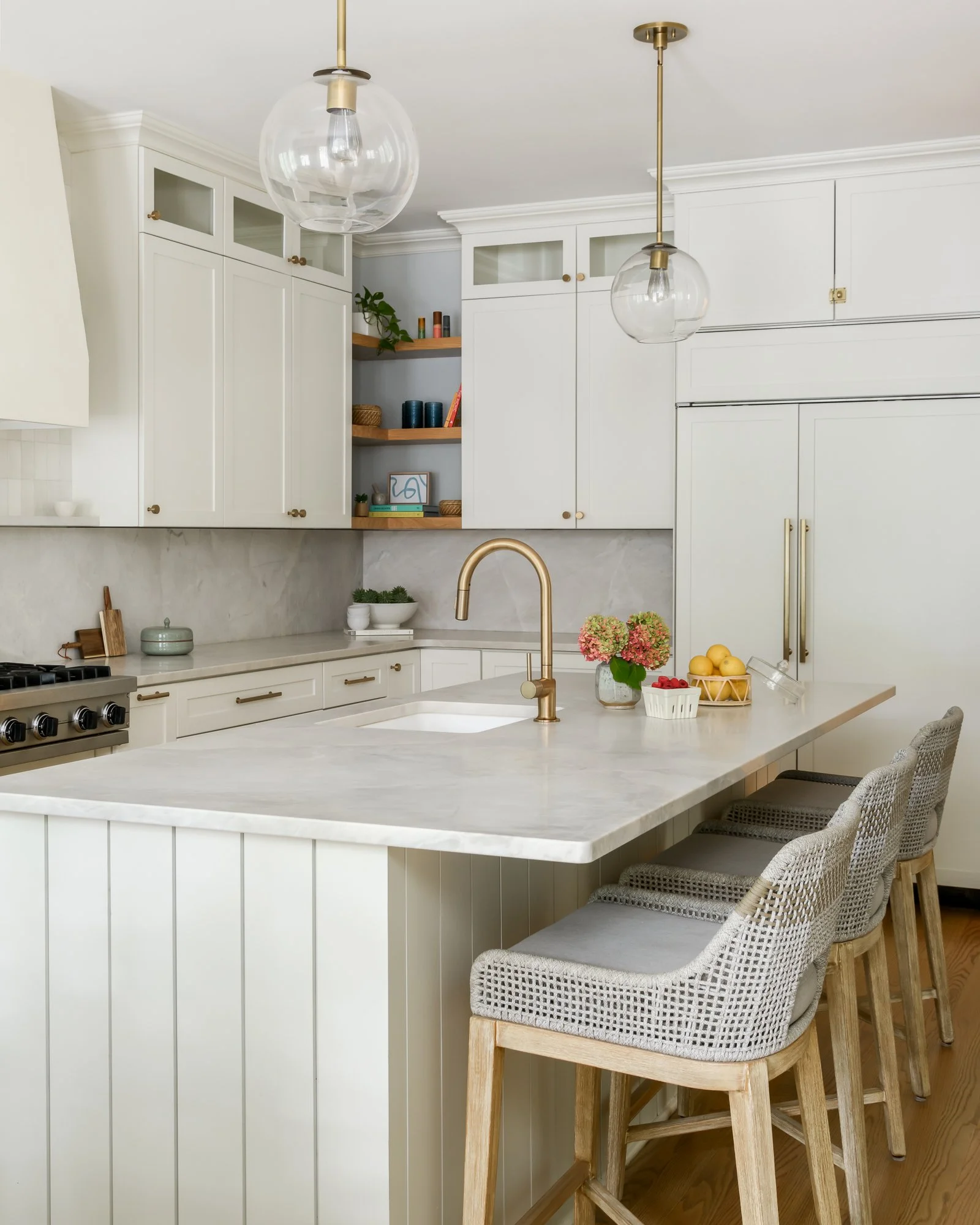 Kitchen with white cabinetry, a large white island with a gold faucet, four chairs with woven gray seats, and decorative items including a vase of flowers and bowls of fruit.