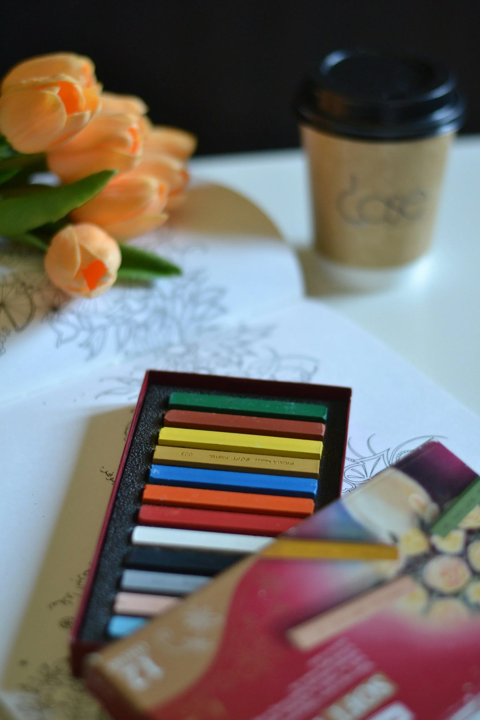 Blurry Foto eines Bechers Kaffee und eines Farbkastens mit Buntstiften auf einem weißen Tisch, mit Blumen im Hintergrund.