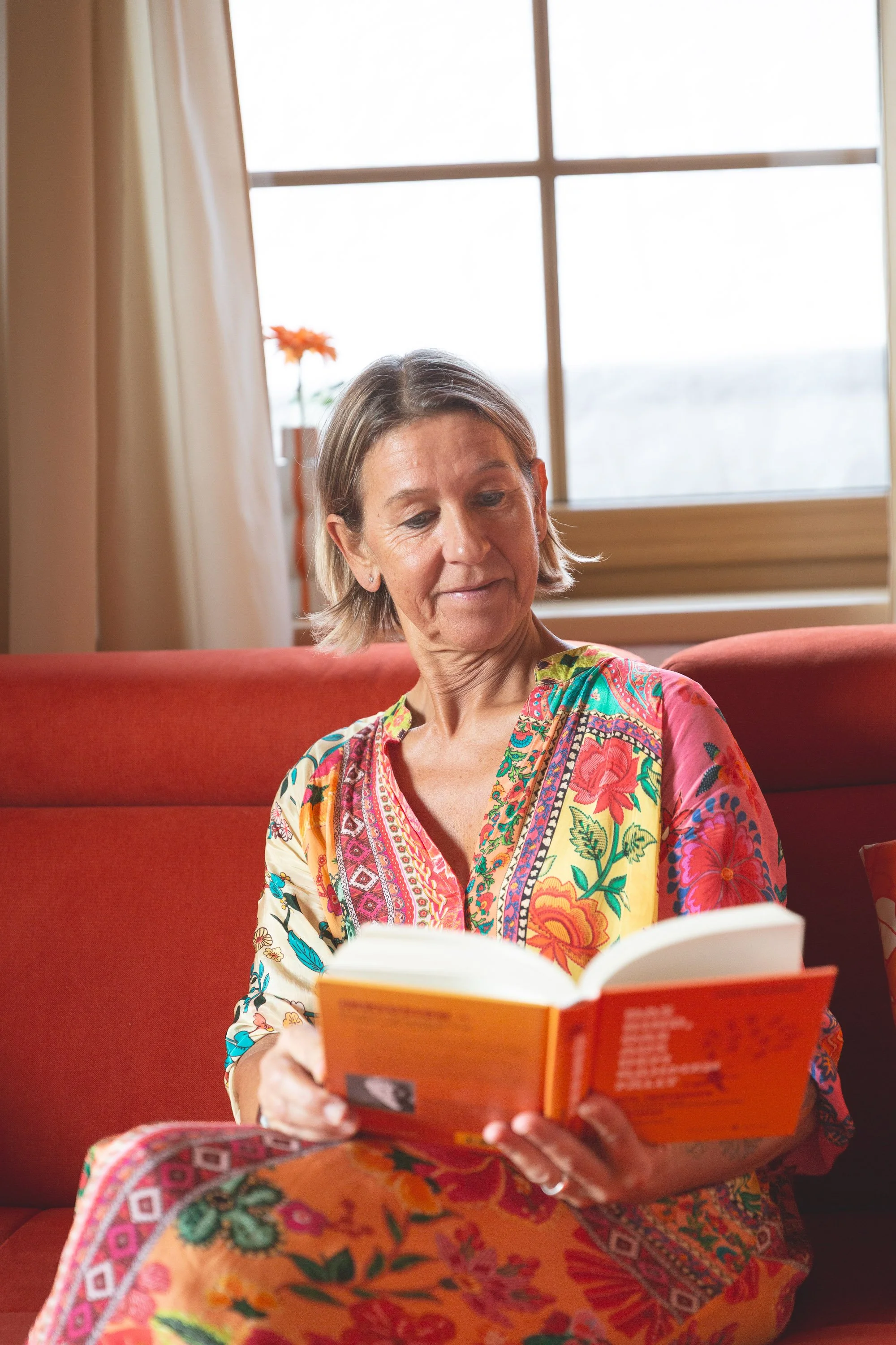 Eine Frau sitzt auf einem roten Sofa, liest ein offenens Buch und trägt ein buntes, florales Kleid. Im Hintergrund befindet sich ein Fenster mit Vorhängen und einer Topfpflanze.