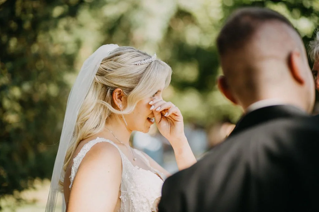 Eine braut trägt ein weißes Kleid und einen Schleier, sie ist emotional während einer Zeremonie umgeben von Bäumen.