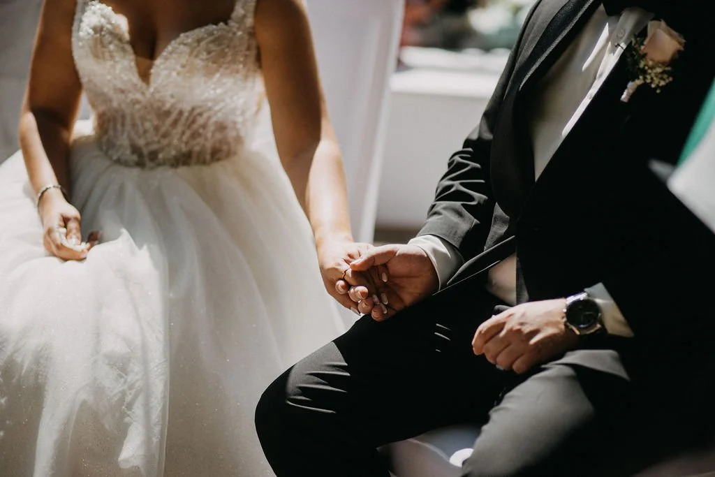Ein Brautpaar hält sich bei der standesamtlichen Trauung an den Händen. Die Braut trägt ein weißes, trägerloses Kleid mit Perl- und Paillettenverzierung. Der Bräutigam trägt einen schwarzen Anzug mit weißem Hemd und Krawatte.