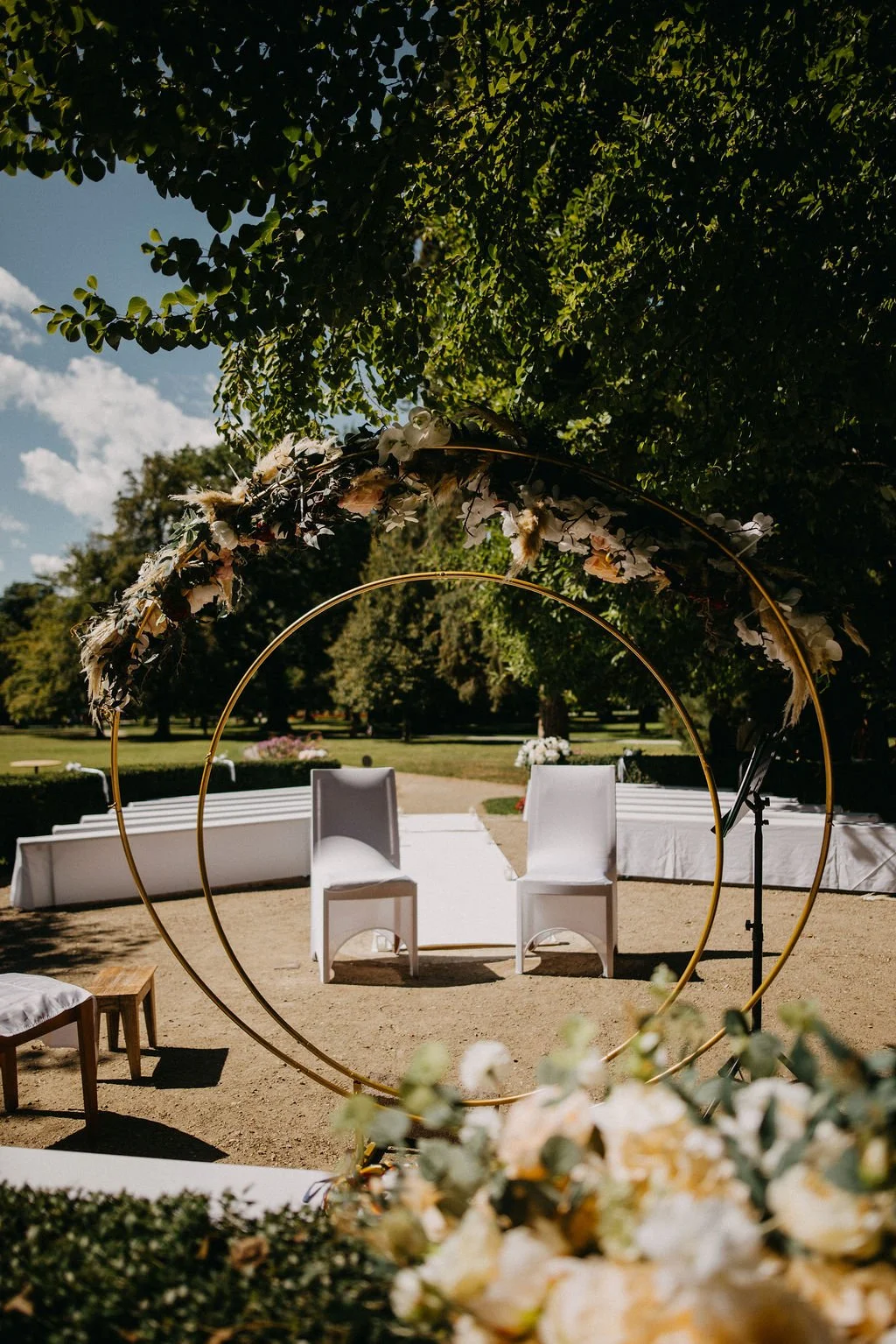 Hochzeitsdekoration im Freien mit zwei weißen Stühlen und goldenen Ringen, geschmückt mit Blumen, unter einem Baum auf einer Gartenwiese.