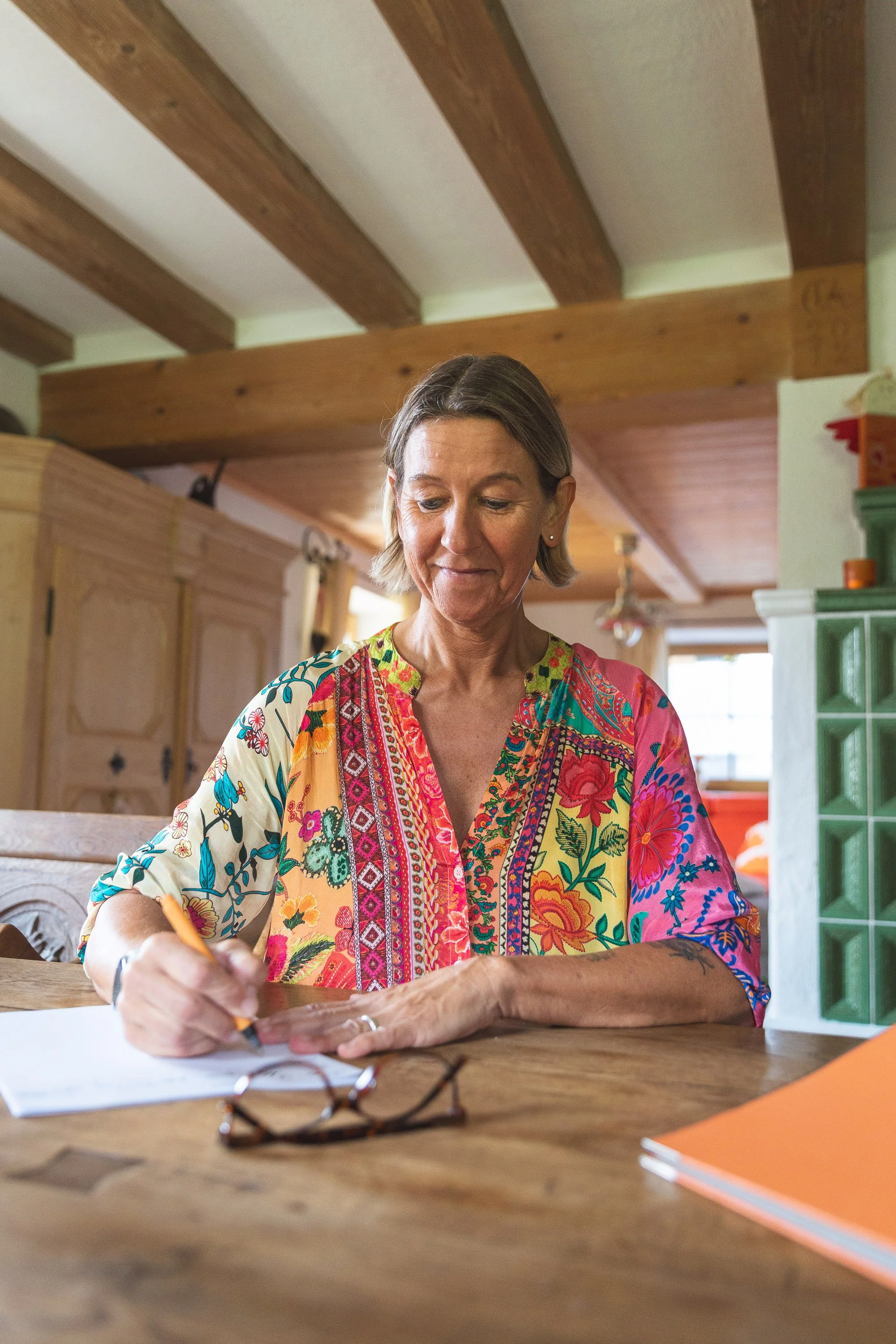 Eine ältere Frau mit kurzen braunen Haaren, die ein buntes, flowerreiches Kleid trägt, sitzt an einem Holztisch und schreibt auf Papier.