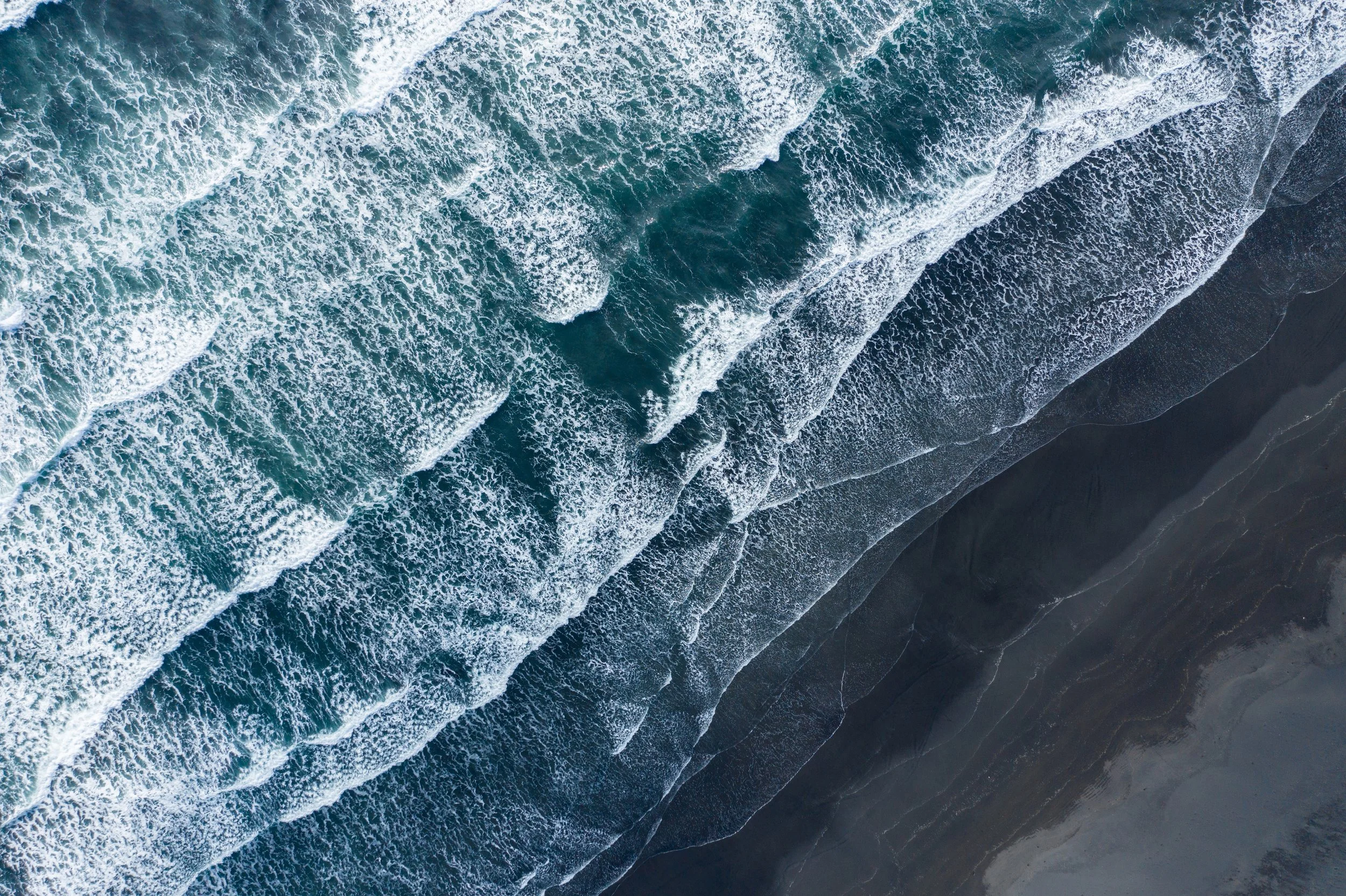 Aerial view of the ocean with waves crashing onto a dark sandy beach.