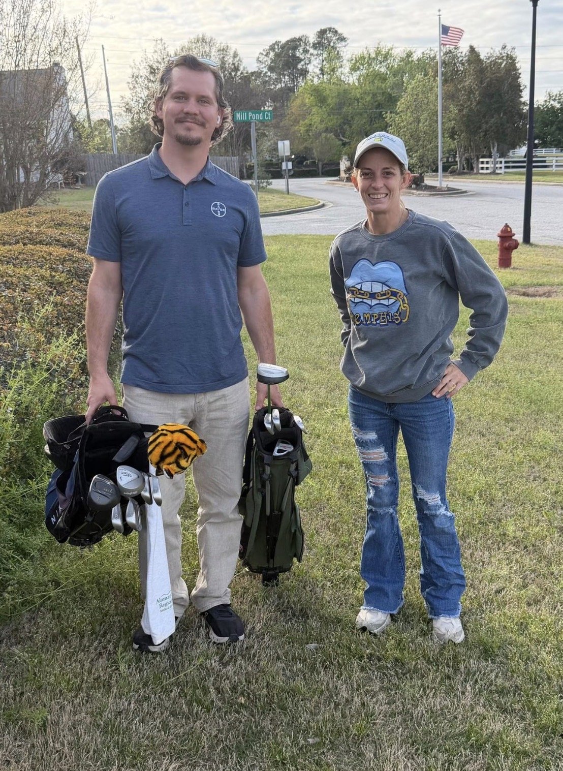 A man and a woman standing on a grassy area, holding golf clubs and golf bags, with a street sign reading 'Mill Pond Ct' behind them, and an American flag flying nearby.