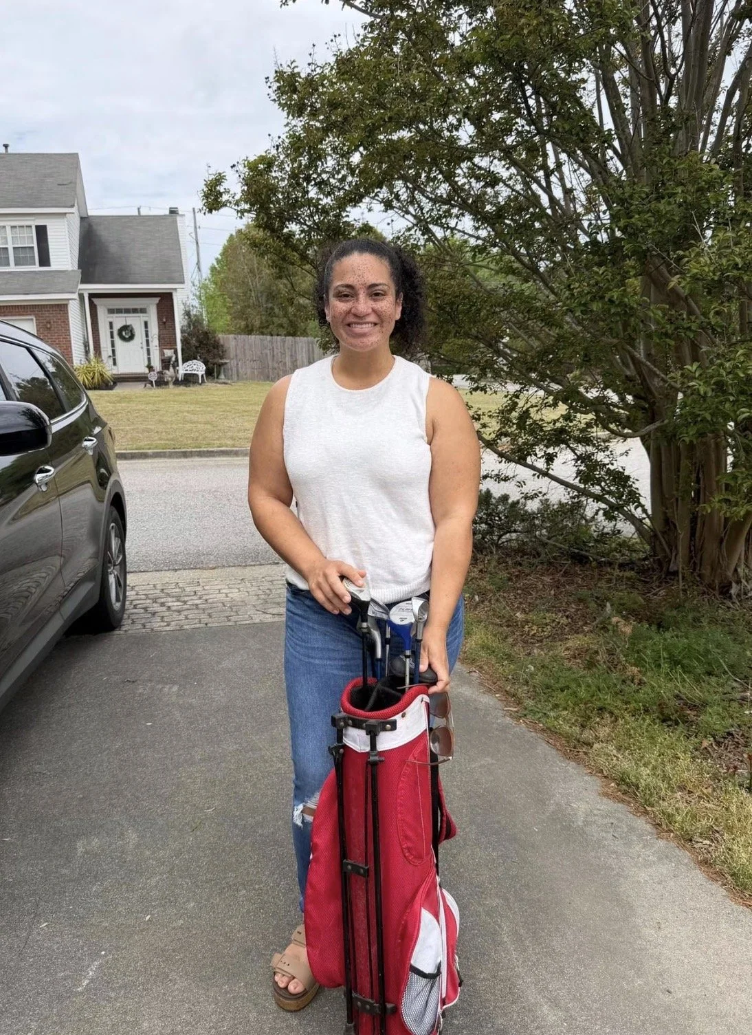 A woman standing outdoors next to a car, holding a golf bag with clubs, smiling, wearing a white sleeveless top and blue jeans.