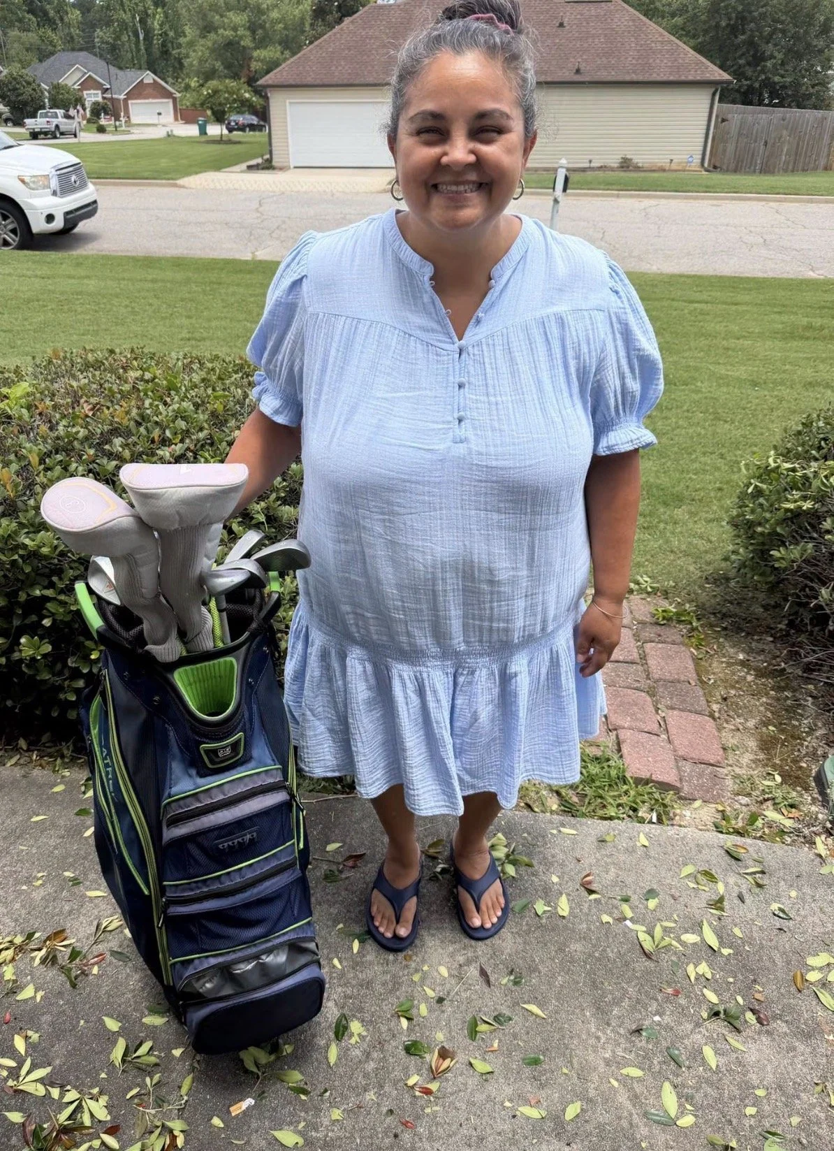 Smiling woman in a light blue dress standing outdoors on a sidewalk with a golf club bag, holding golf clubs, and surrounded by a manicured lawn and houses in the background.