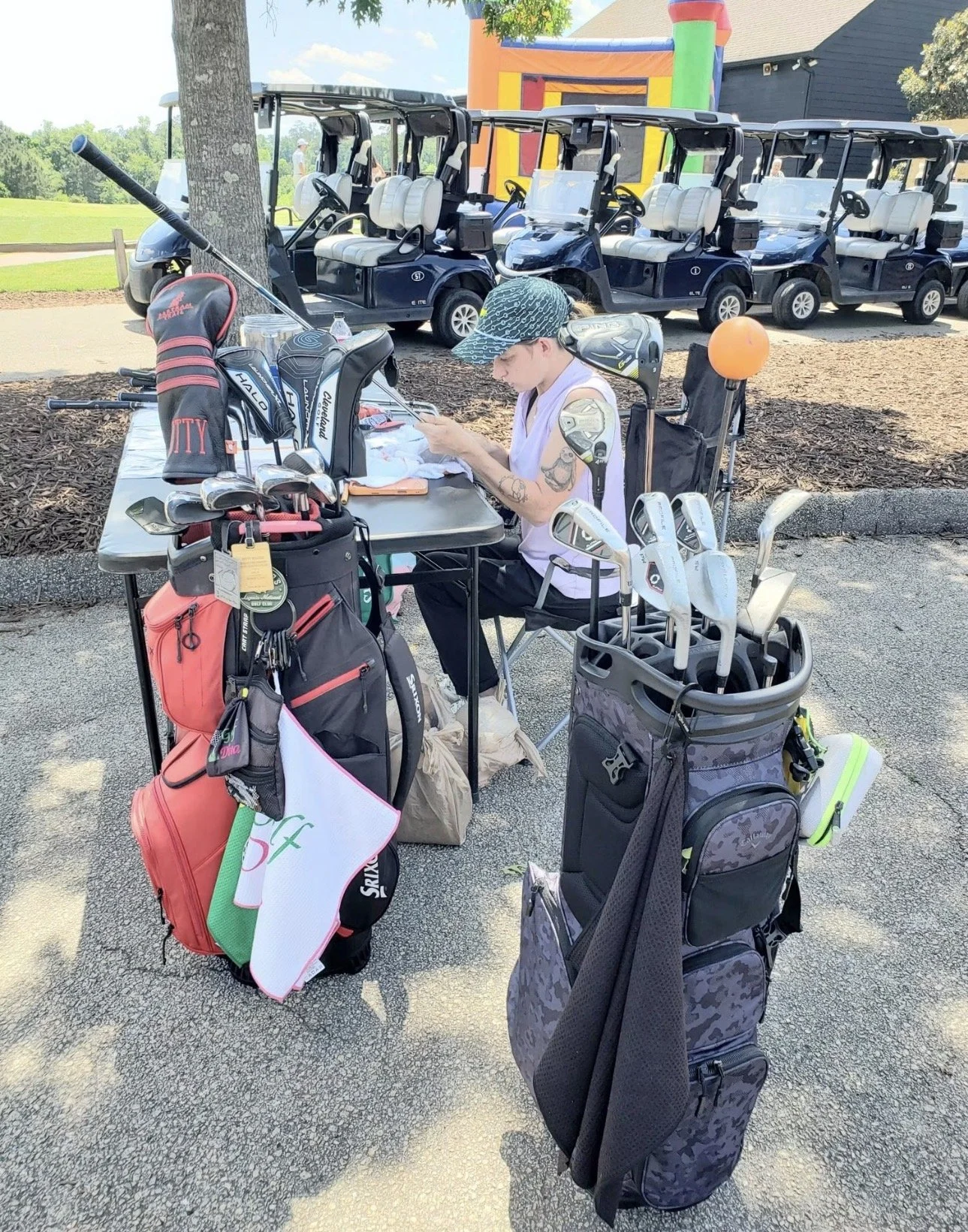 A woman sitting at a table outdoors surrounded by golf clubs, bags, and equipment, with a line of golf carts and a colorful inflatable in the background at a golf course.
