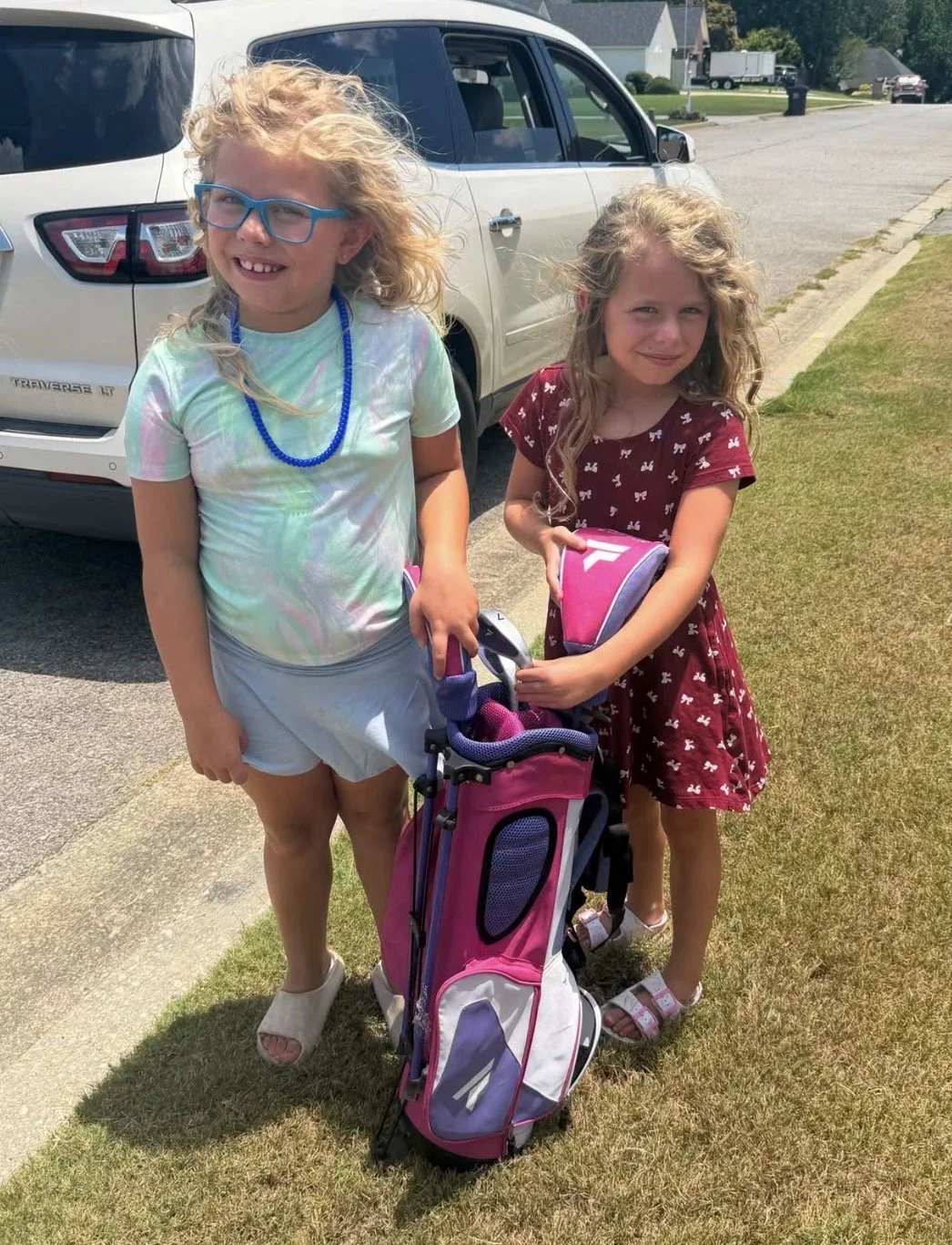 Two young girls stand outdoors on a sidewalk next to a parked white SUV, with a golf bag and a pink golf club set between them. One girl has blond curly hair, wears blue glasses, a colorful tie-dye shirt, light shorts, and sandals. The other girl has curly hair, wears a maroon dress with white butterfly patterns and sandals. They are smiling and squinting in the sunlight.