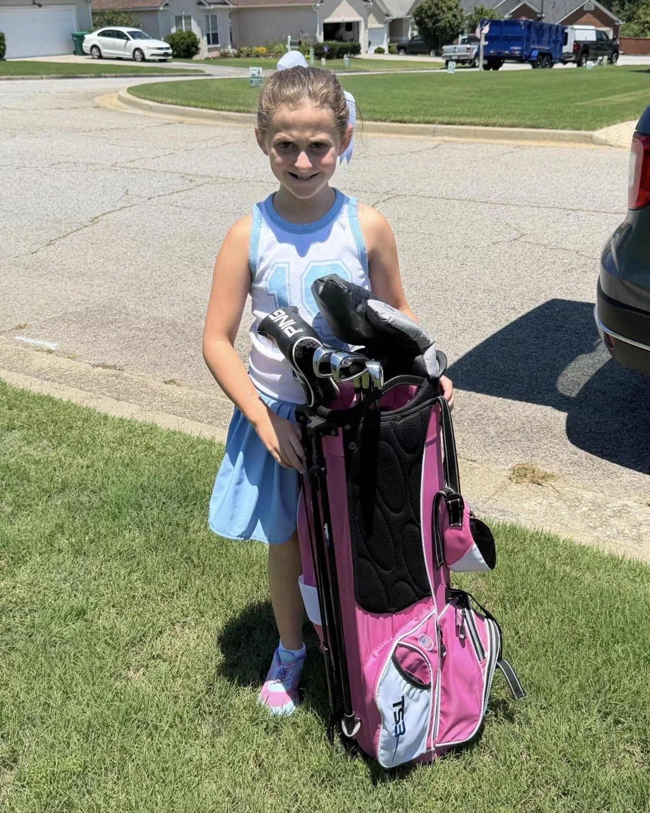 A young girl with a big smile wearing a white and blue sports outfit, standing outdoors on grass next to a pink golf bag with clubs.