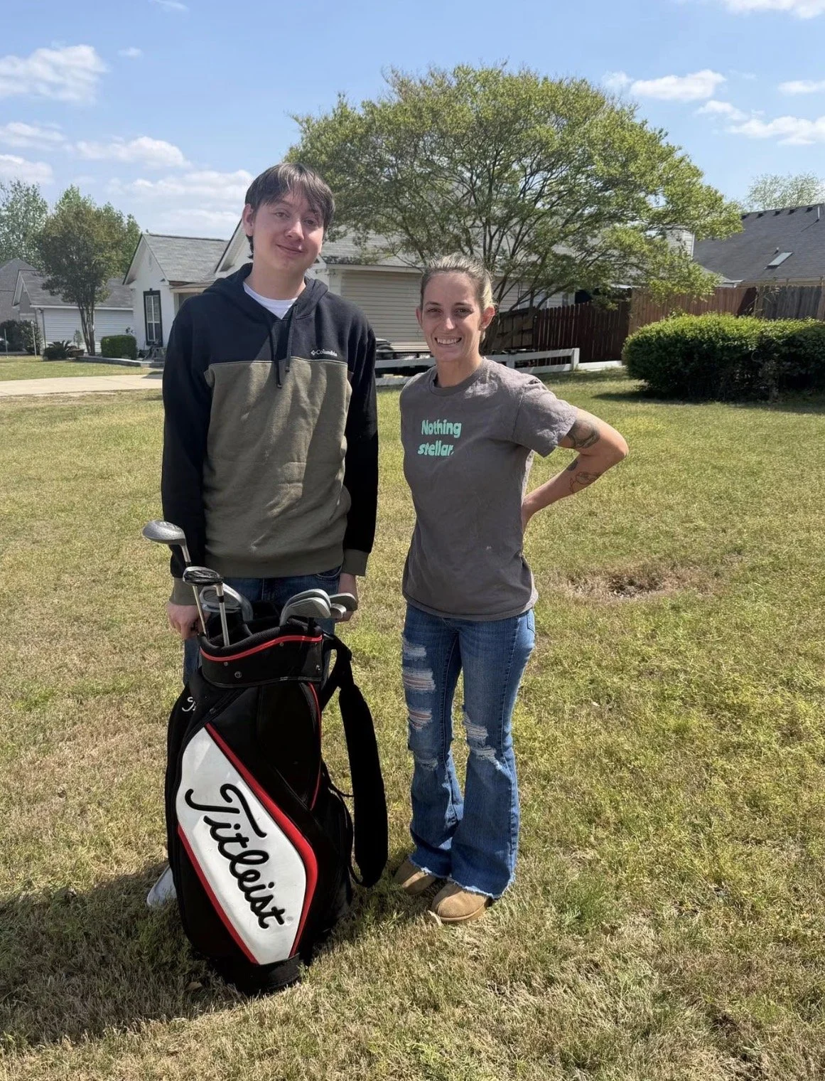 A young man and a woman standing on a grassy yard outdoors on a sunny day. The young man is holding a golf club and has a golf bag with clubs next to him. The woman is smiling and wearing a T-shirt that says 'Nothing stellar'. There are houses, trees, and a blue sky in the background.
