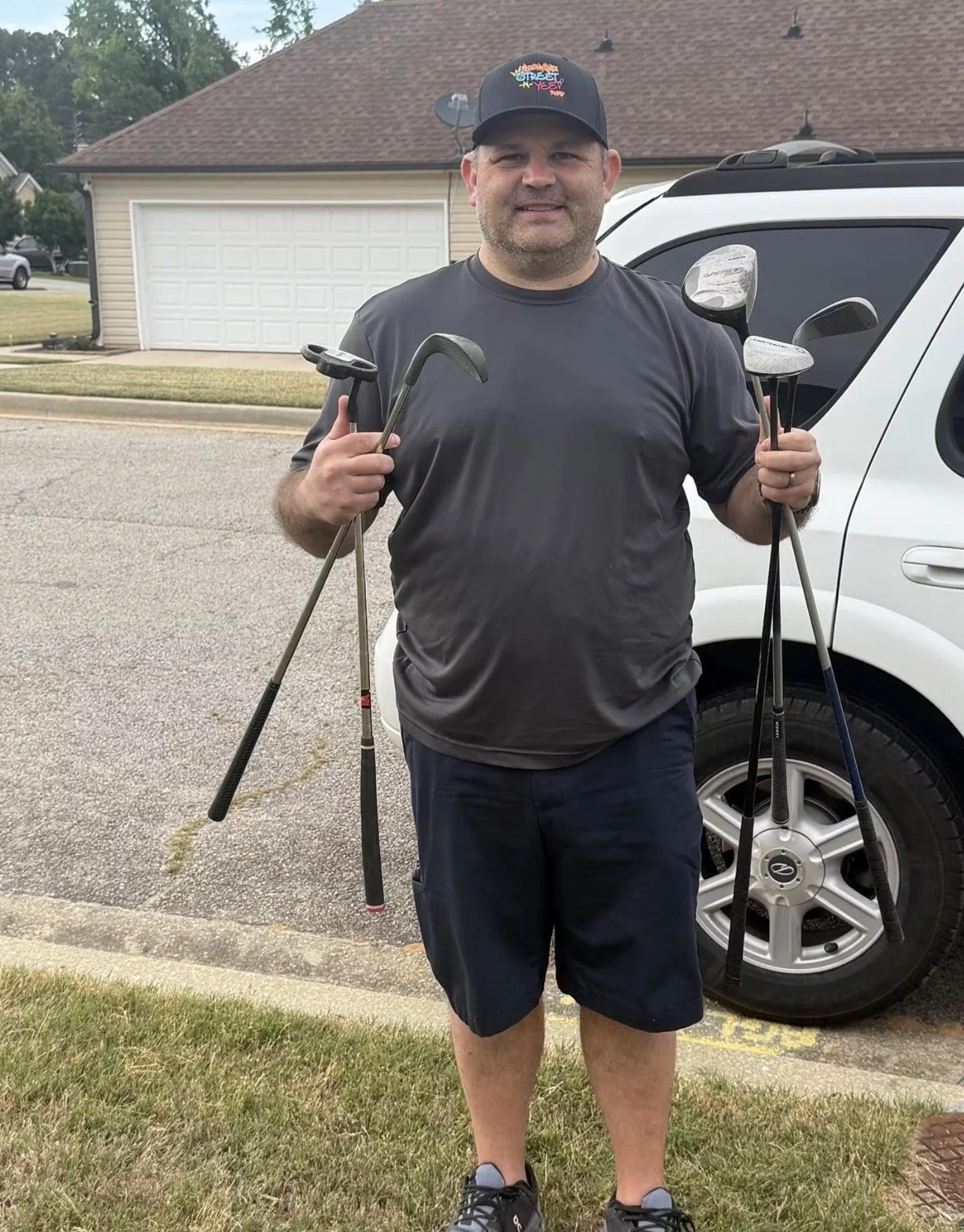 A man standing outdoors near a white vehicle, holding several golf clubs, wearing a black cap, a dark gray t-shirt, black shorts, and athletic shoes.