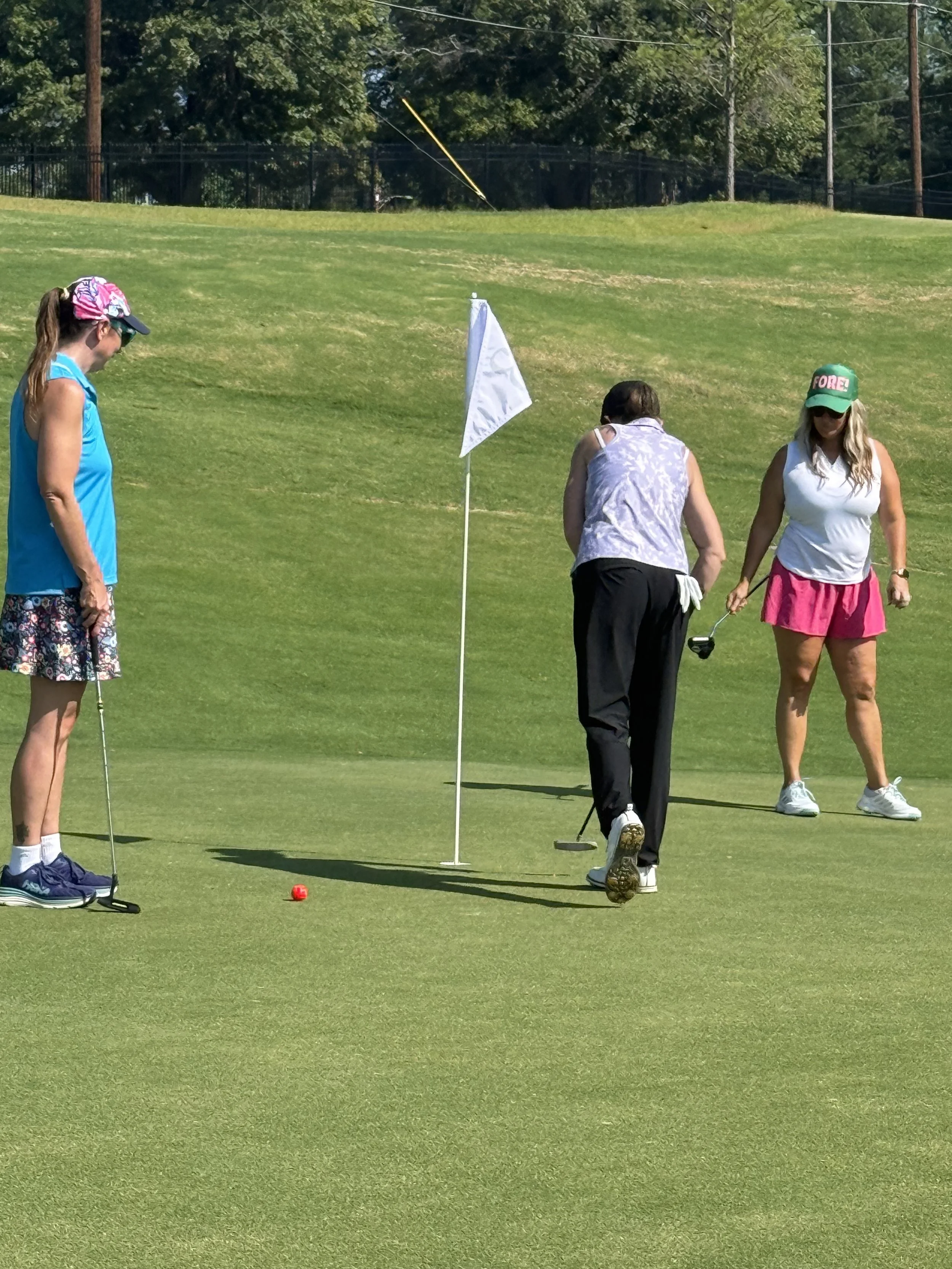 Three women on a golf course near a flag, with a golf ball on the grass and a caddy holding clubs.