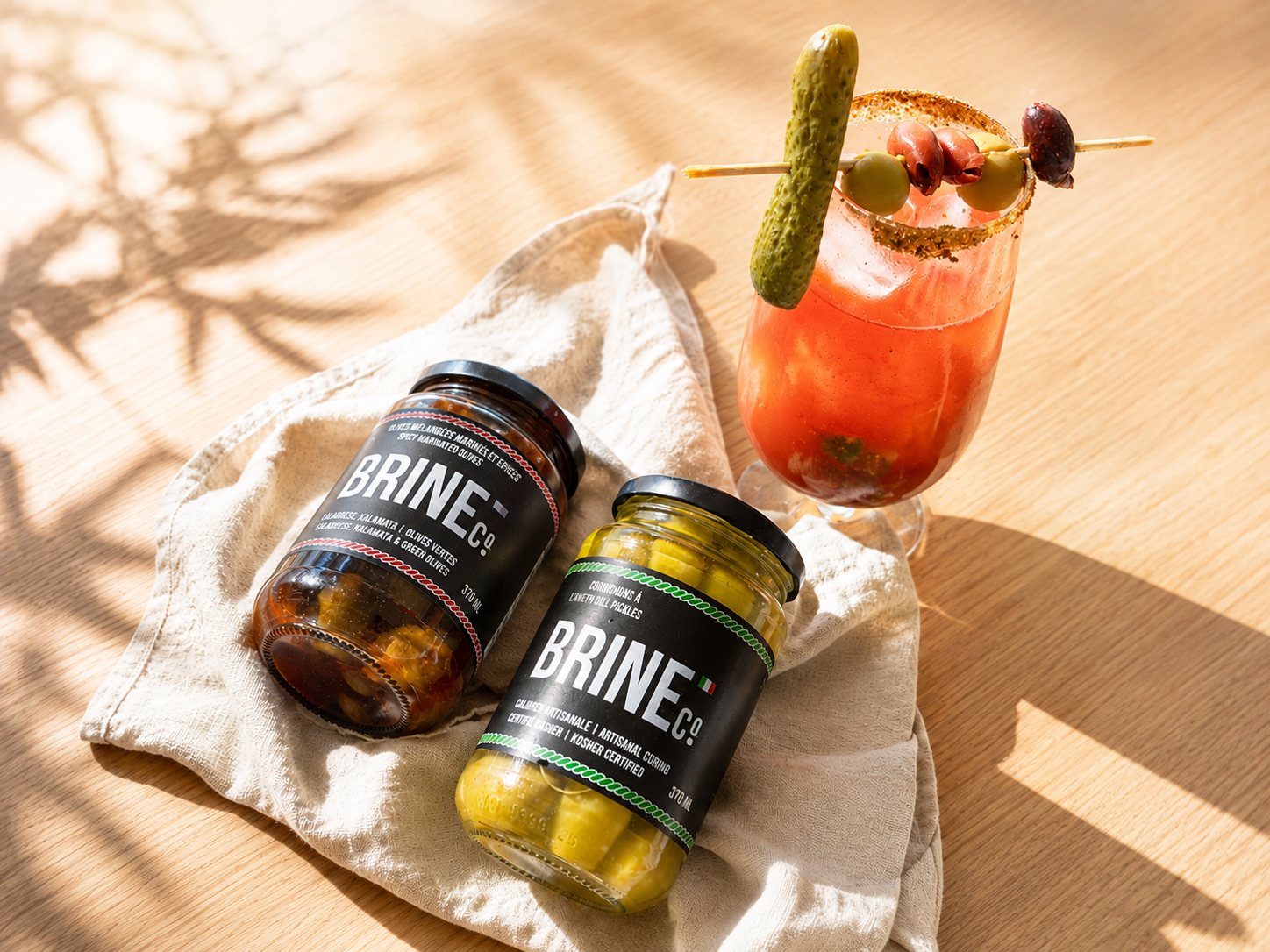 A cocktail garnished with a pickle and olives, alongside two jars of Brine Co. pickled vegetables on a cloth and wooden surface with shadows.