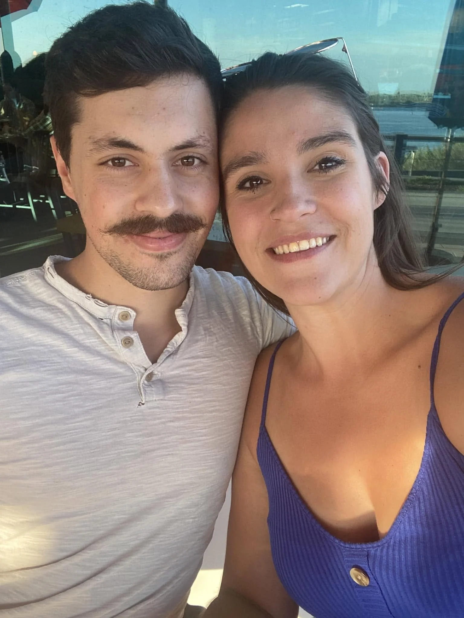 Close-up photo of a smiling man and woman taking a selfie together indoors with large windows behind them, showing outdoor scenery and reflections.