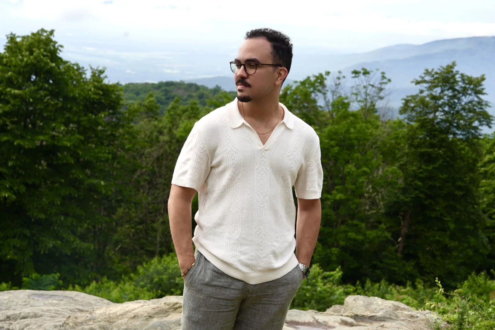 Un hombre con gafas, barba, vestido con suéter blanco y pantalones grises, de pie en un entorno natural con árboles y montañas de fondo.