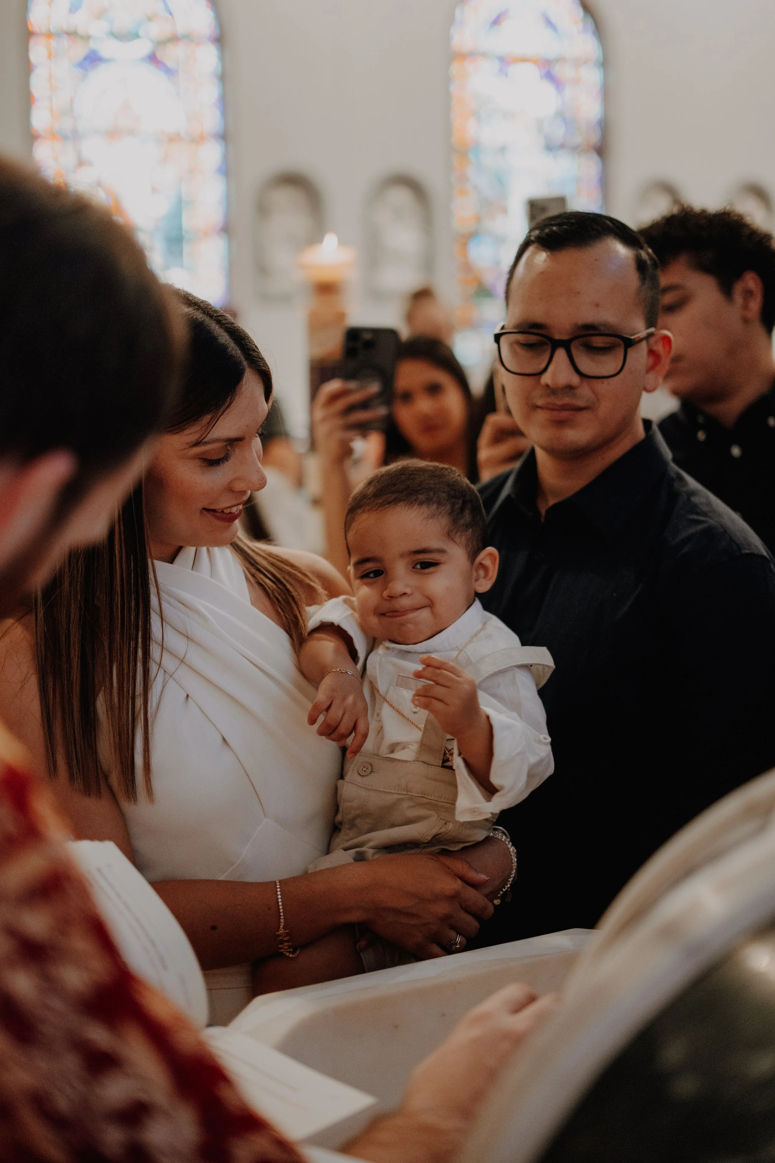 Personas participando en una ceremonia en una iglesia, con ventanas de vitral en el fondo, una mujer sostiene a un niño y otros adultos toman fotos.