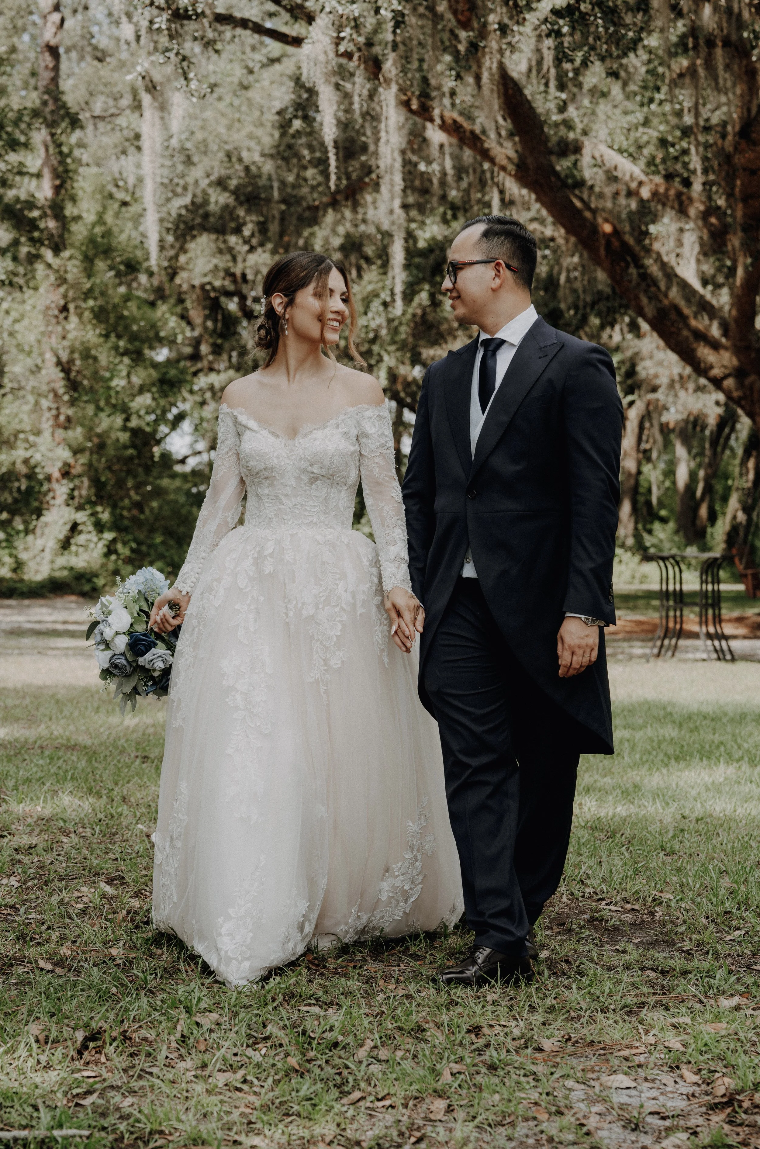 Pareja de recién casados caminando en un parque, la mujer lleva vestido de novia y el hombre traje oscuro, ambos sonrientes y tomados de la mano.