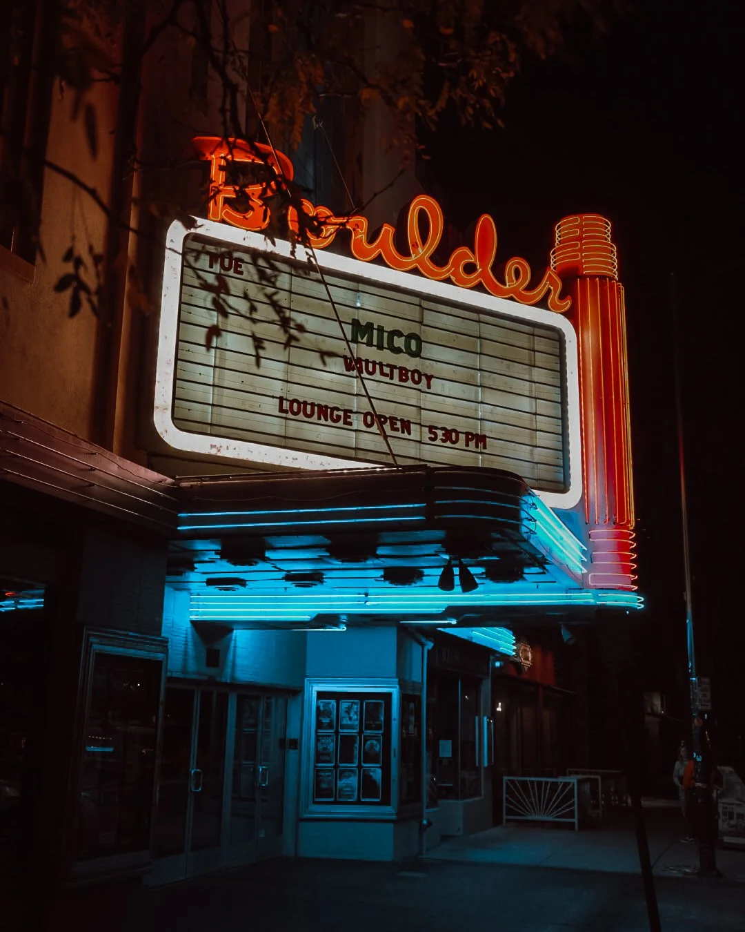 Cartel luminoso en un cine llamado 'Cine Crueldad' con letras en neón rojas y azul, exterior de ingreso con puertas de vidrio y cartelera de películas visible.