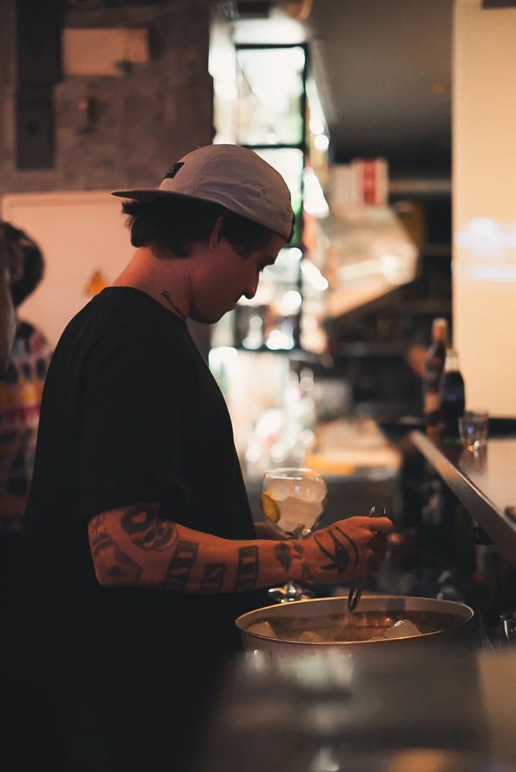 Joven con sombrero gris y tatuajes en el brazo, preparando una bebida en un bar con fondo desenfocado y luz cálida.