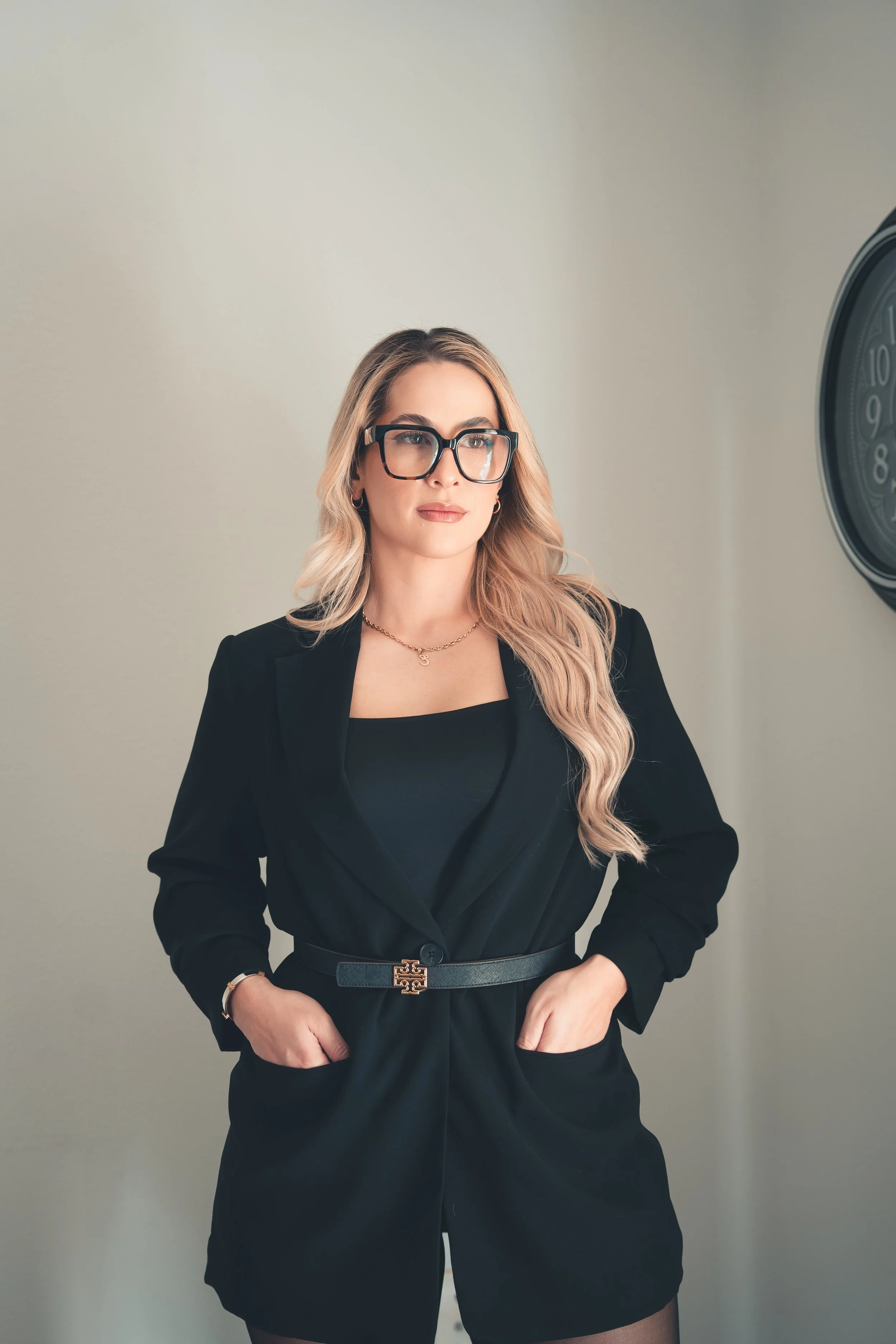 Mujer con cabello rubio, gafas grandes, usando traje negro con cinturón dorado y accesorios, posando en un fondo neutro, cerca de un reloj grande en la pared.