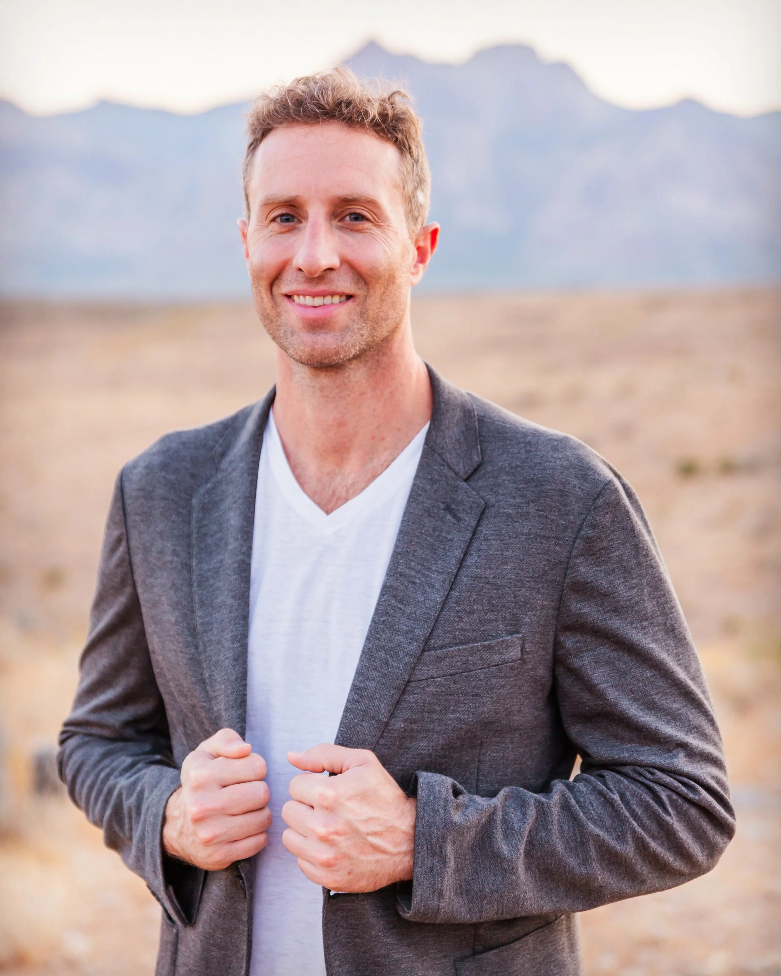 A smiling man in a gray blazer and white shirt standing outdoors in a desert with mountains in the background.