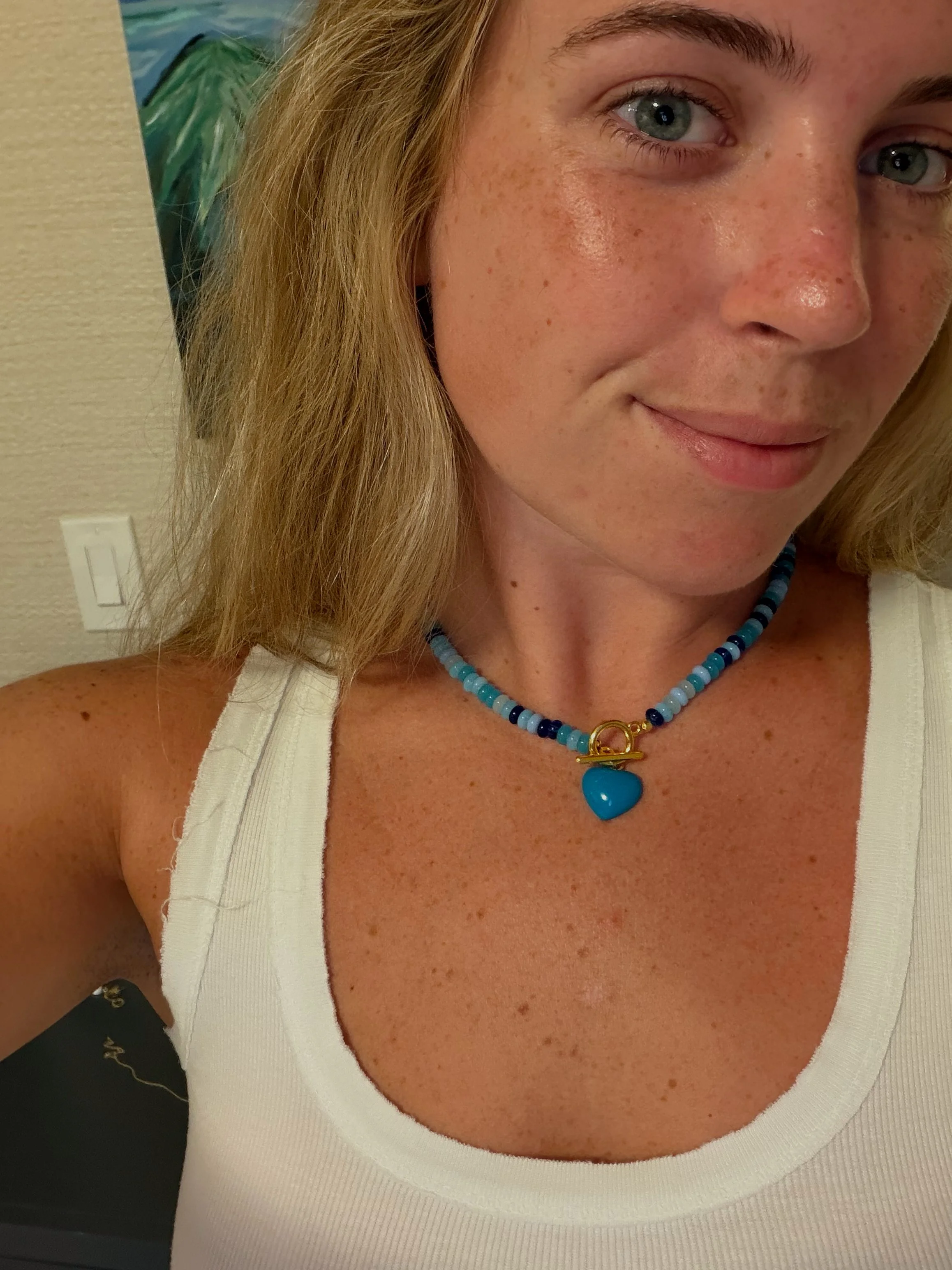 Close-up of a woman's face, showing her eye, cheek, and part of her nose. She has blonde hair, blue eyes, and freckles. She wears a white top and a blue beaded necklace with a blue heart pendant.