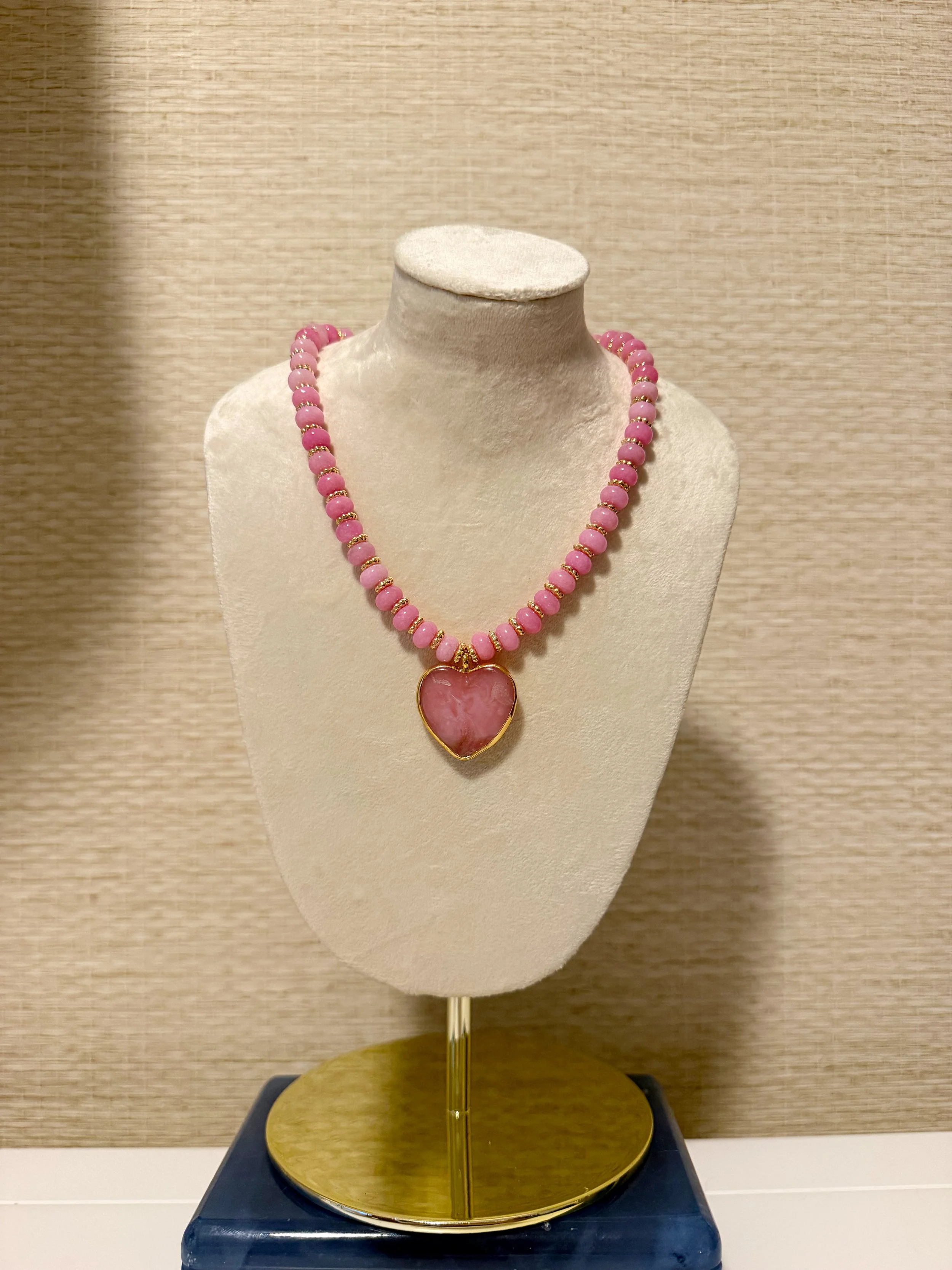 Pink beaded necklace with a large heart-shaped pendant displayed on a beige jewelry bust on a podium.