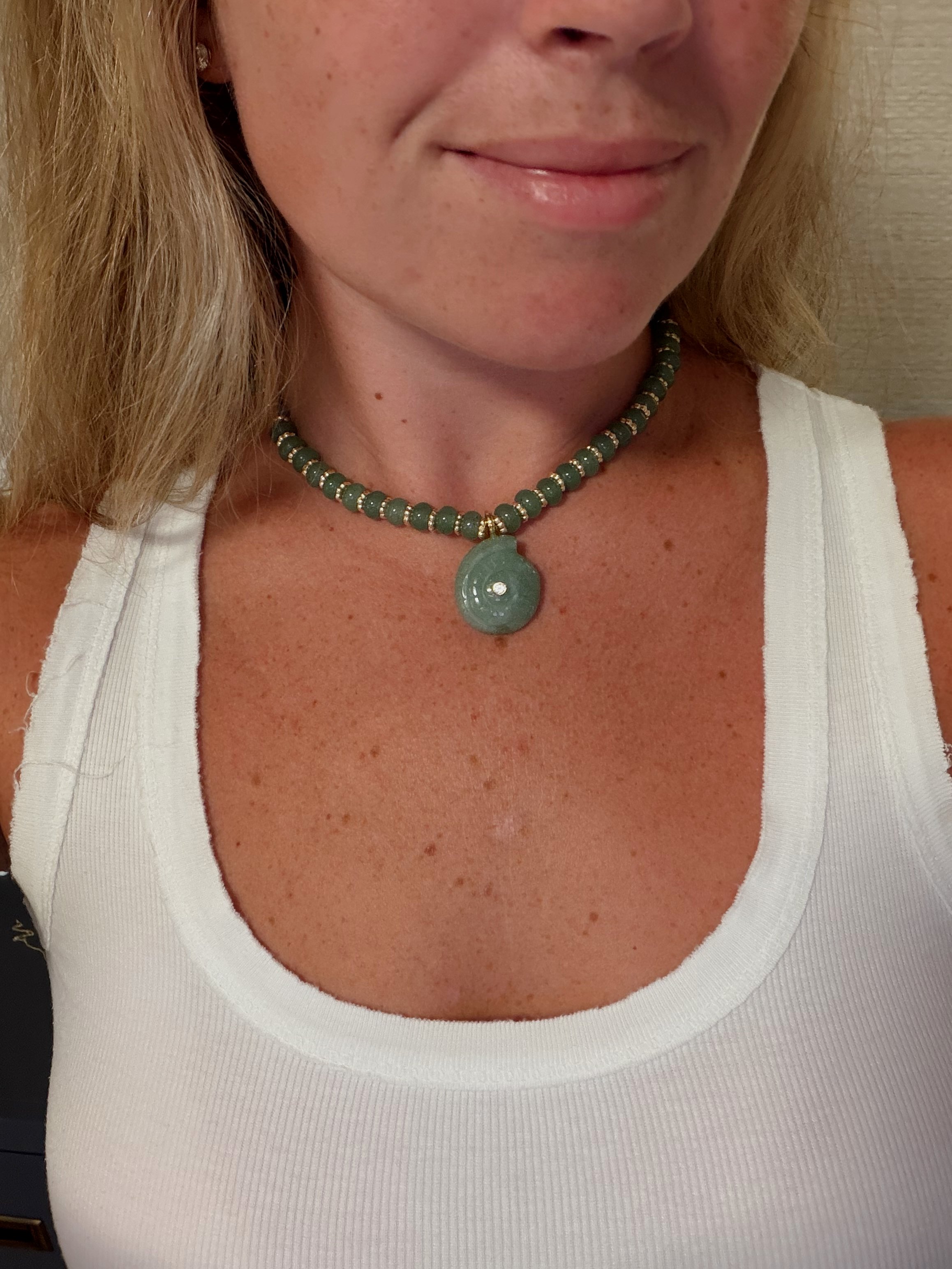 A woman wearing a white tank top and a green beaded necklace with a circular pendant.