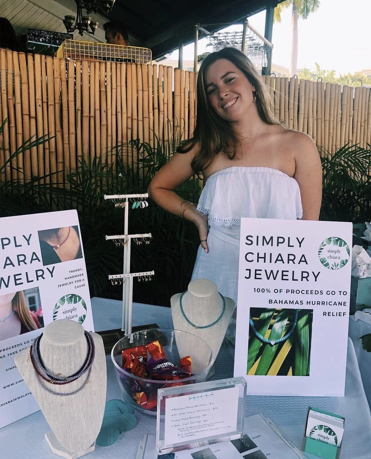 Woman smiling behind a jewelry display table with necklaces, signs, and jewelry pieces for sale at an outdoor market, with bamboo fencing and greenery in the background.