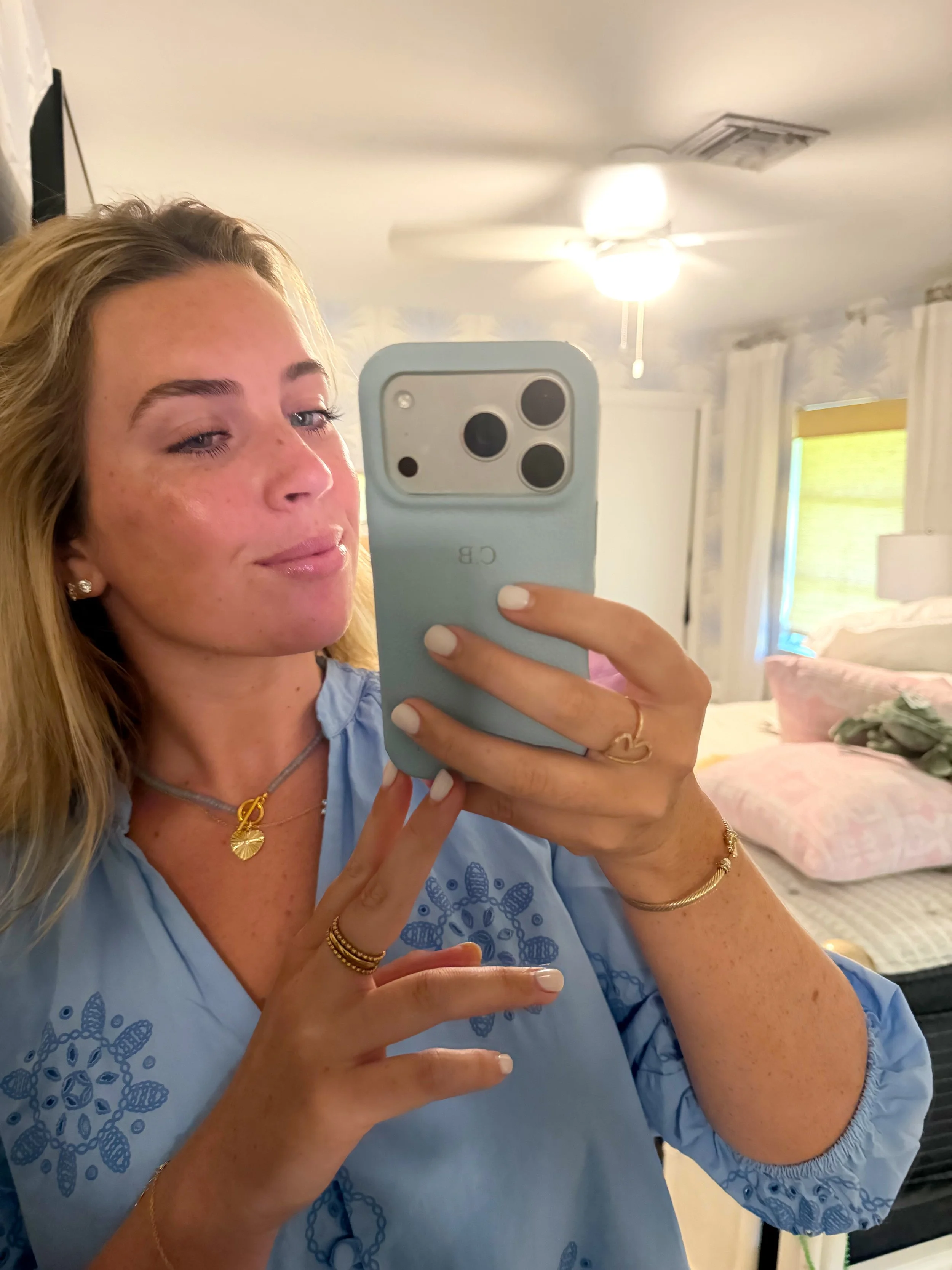 A woman with blonde hair taking a mirror selfie with an iPhone in a bedroom. She is wearing a blue embroidered top, jewelry, and has a slight smile. The background shows a bed with pink pillows and a window with yellow curtains.