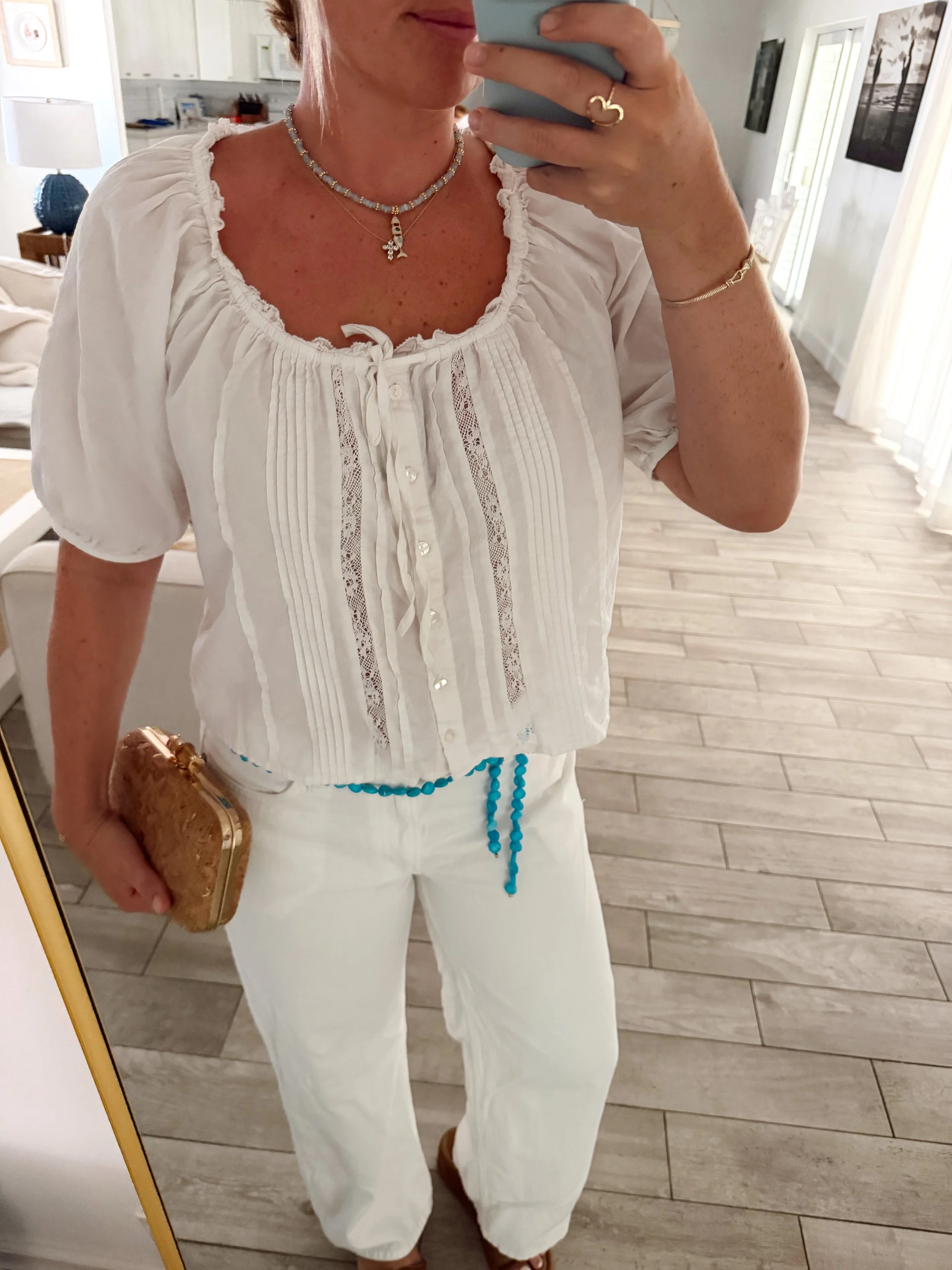 A woman taking a mirror selfie, showing her white ruffled blouse with a button-down front, white pants with a blue bead necklace, jewelry including a necklace with a cross pendant, a bracelet, and a ring, holding a clutch bag in her left hand in a room with wooden flooring, a stand lamp, and framed pictures on the wall.
