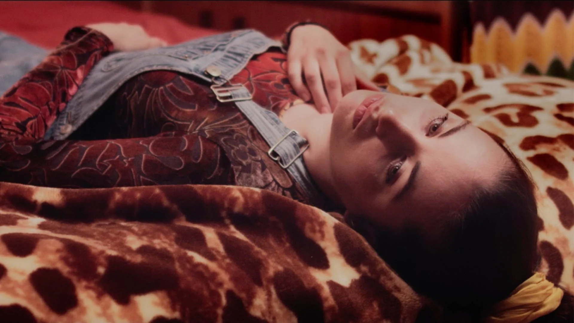 Jeune femme allongée sur un lit avec couvertures à motif léopard, portant une salopette en jean et un haut à motifs floraux, regardant vers l'objectif.