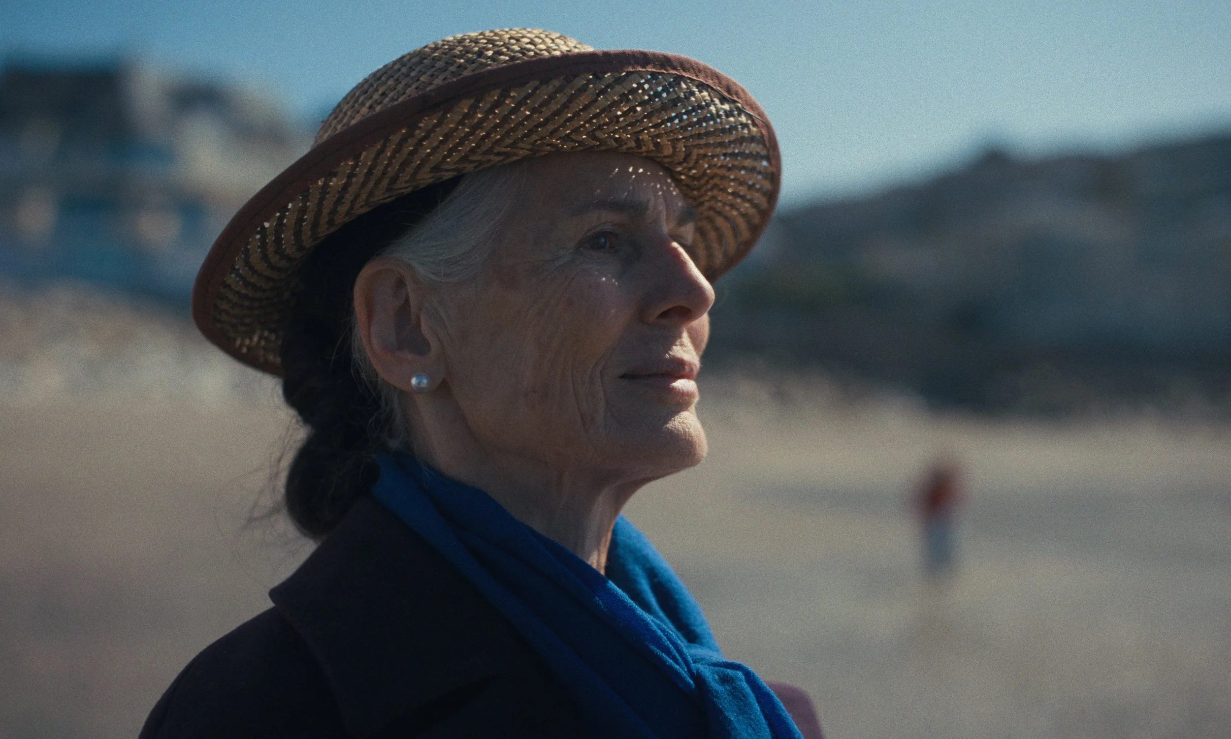 Une femme âgée portant un chapeau en paille regarde vers la droite, en arrière-plan une plage de Normandie.