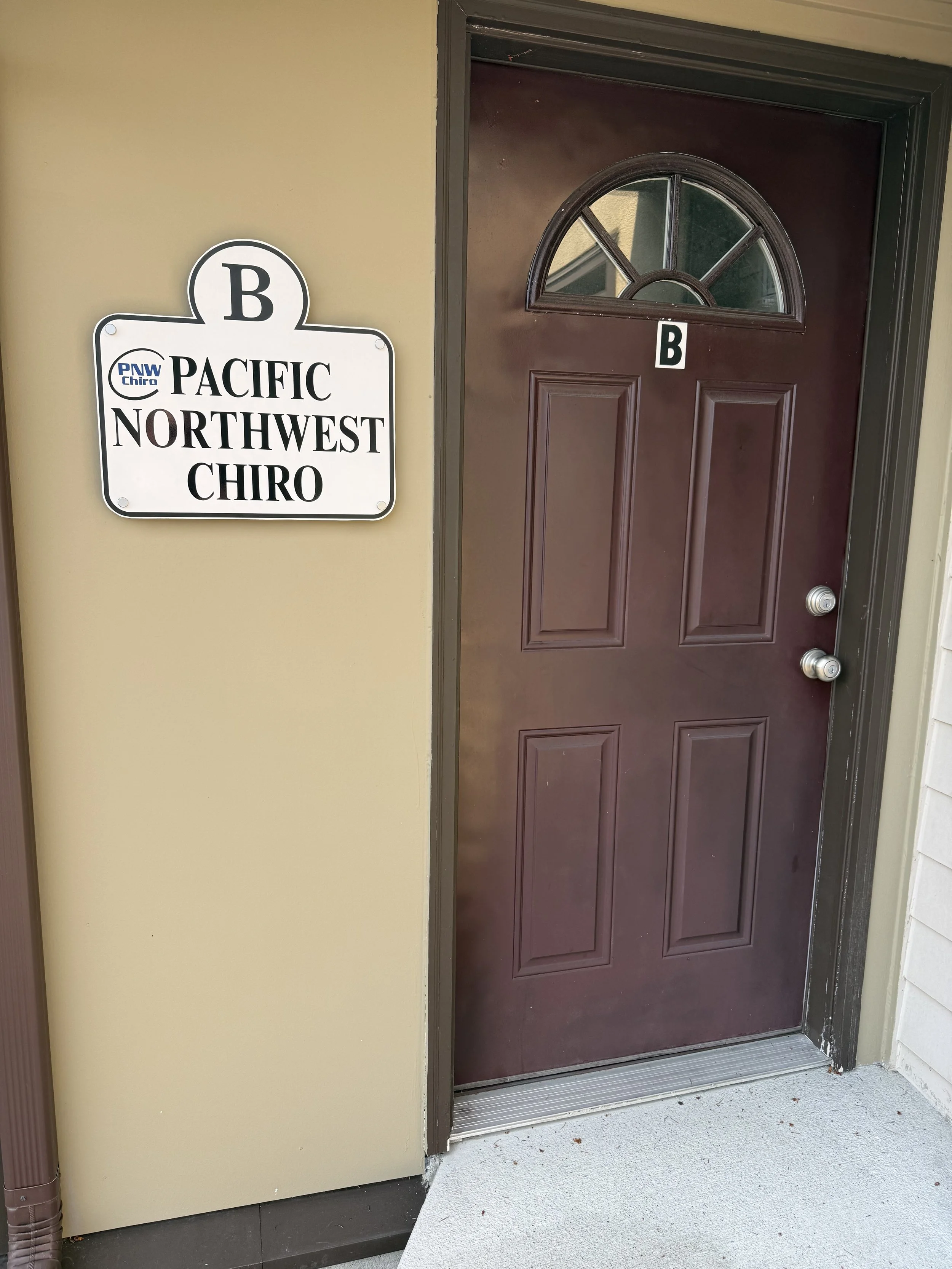 A brown door with a half-circle window near the top, and the letter B on a white label in the center of the door. To the left of the door is a sign with the text 'Pacific Northwest Chiro' and a large B at the top.