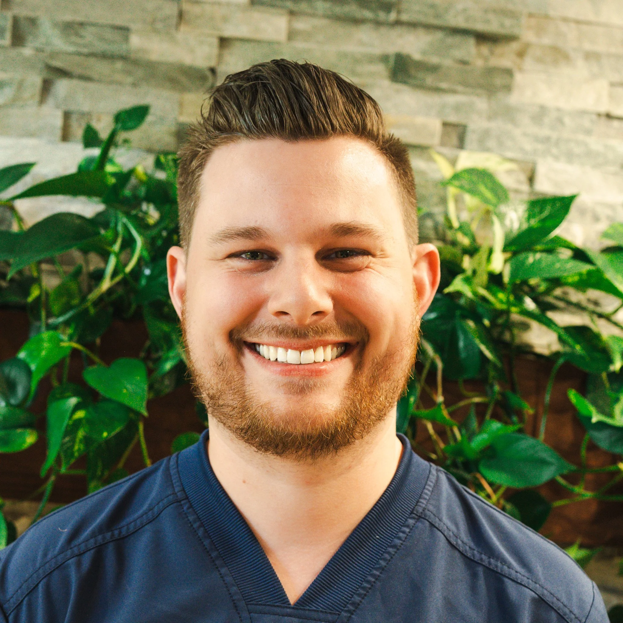Close-up of a smiling man with short, styled hair, a beard, and mustache, wearing a dark blue shirt, standing in front of a wall with green plants.