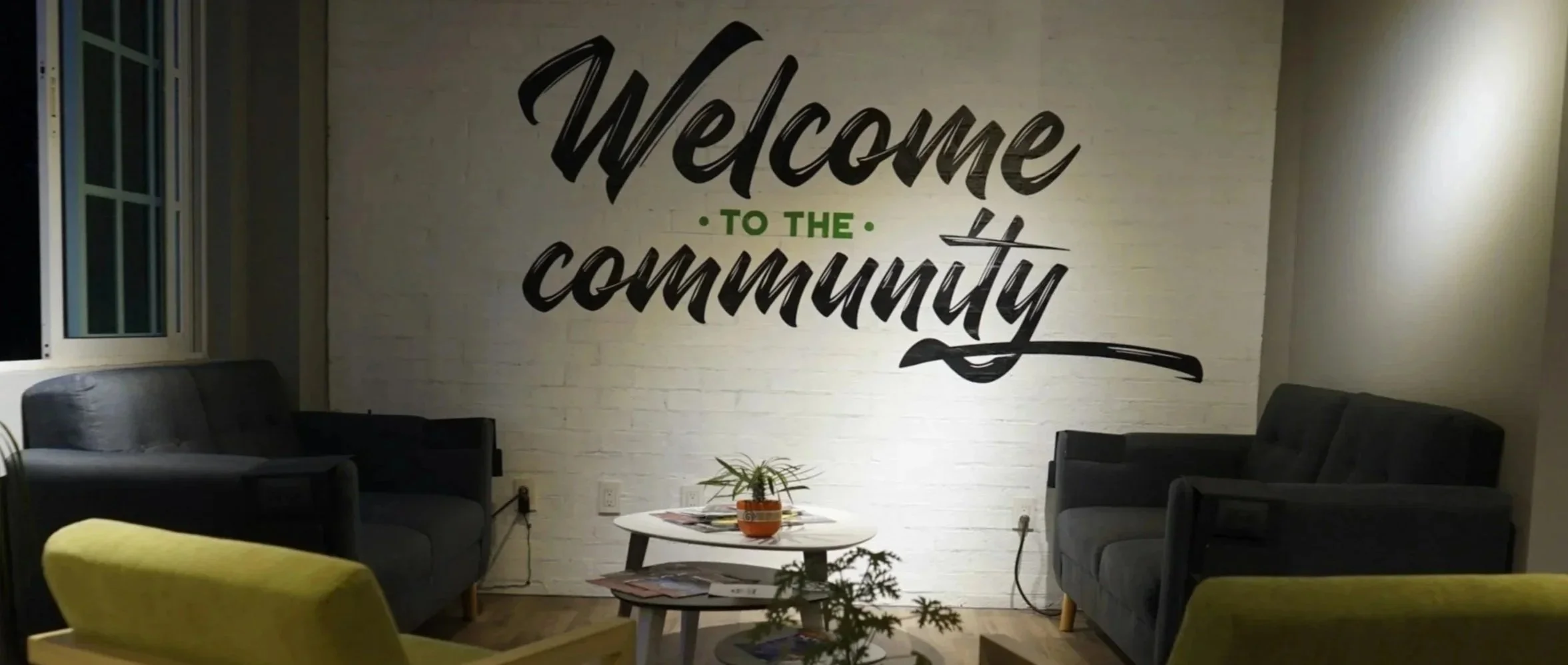 Living room with white brick wall featuring the phrase 'Welcome to the Community' in large black and green lettering, two gray sofas, a small round coffee table with magazines and a potted plant, and a yellow chair in the foreground.