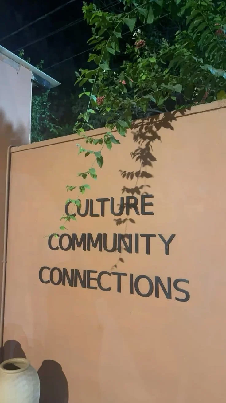 A beige wall with black lettering that reads 'CULTURE COMMUNITY CONNECTIONS.' There are green plants and leaves hanging from above, casting shadows on the wall, with a glimpse of a house roof and dark sky in the background.