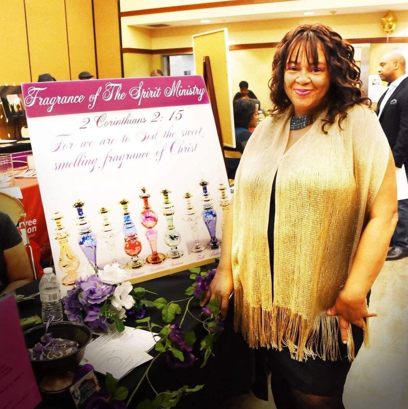 A woman with curly hair wearing a gold-colored shawl standing next to a display board with the title 'Fragrance of The Spirit Ministry' and a quote from 2 Corinthians 2:15. The display features various colorful perfume bottles and purple flowers on the table.