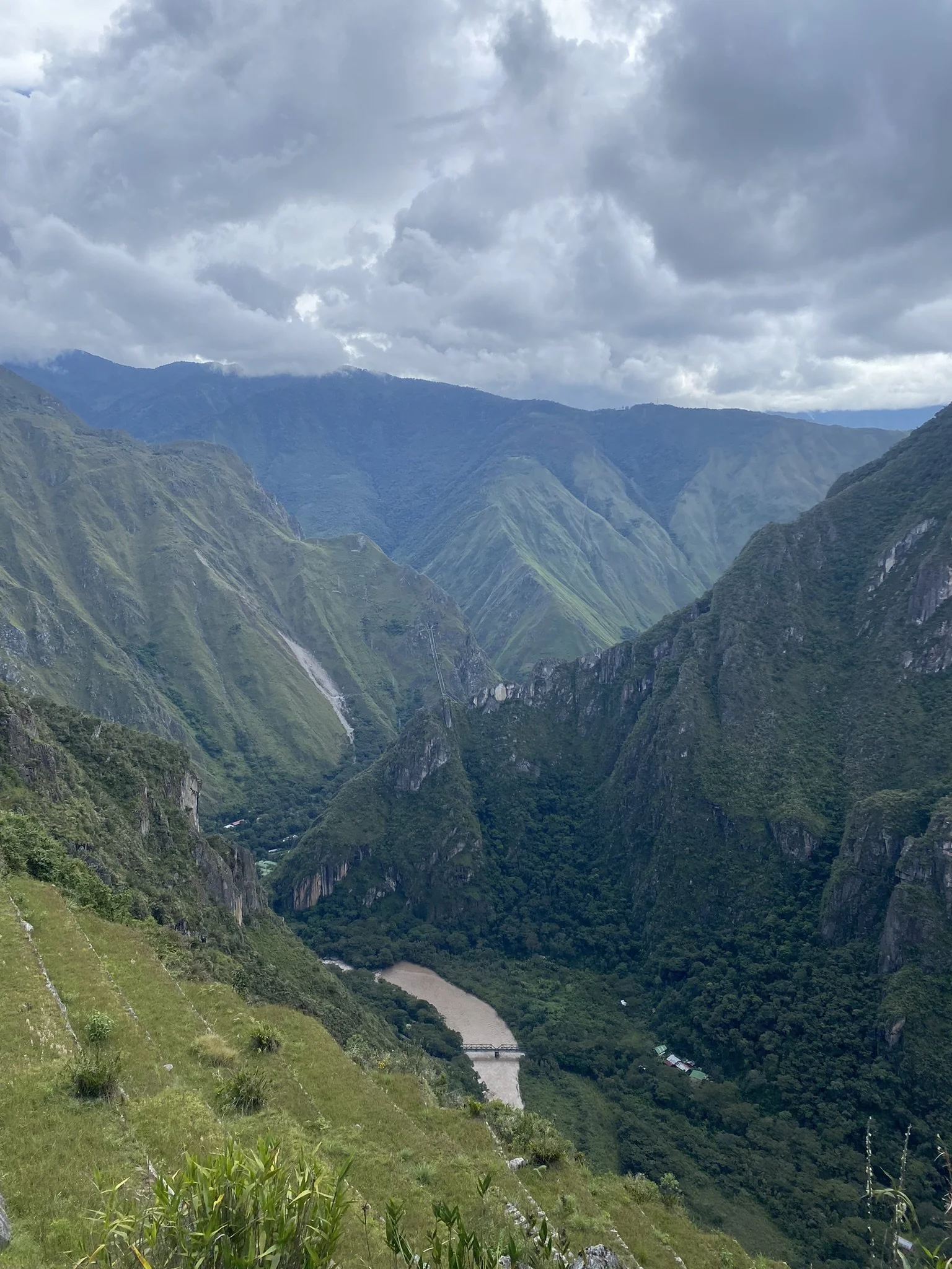A mountain valley with lush green peaks, a river running through the bottom, and a small bridge crossing the river. The sky is cloudy.