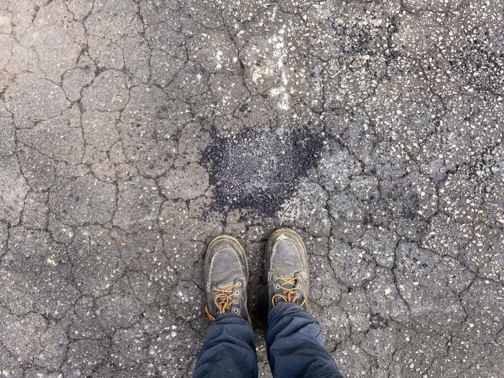 Dark cold-mix asphalt filling a small, irregular pothole after a throw-and-roll patch repair in Durham, NC, surrounded by cracked pavement.
