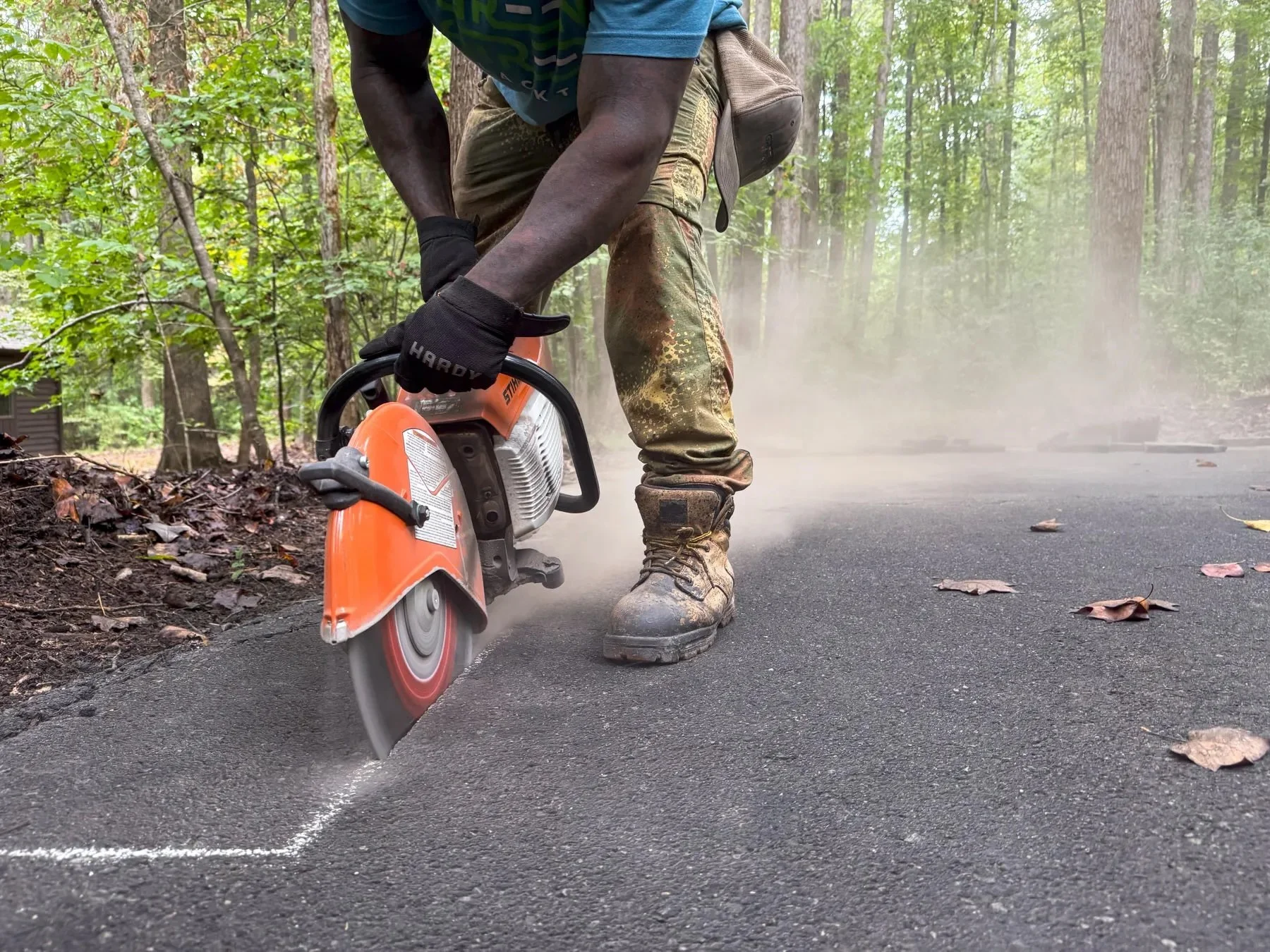 Green Street Blacktop crew member using a handheld cut-off saw to score a clean line into an asphalt driveway ahead of a patch repair, dust rising from the blade, wooded Durham, NC property in the background.