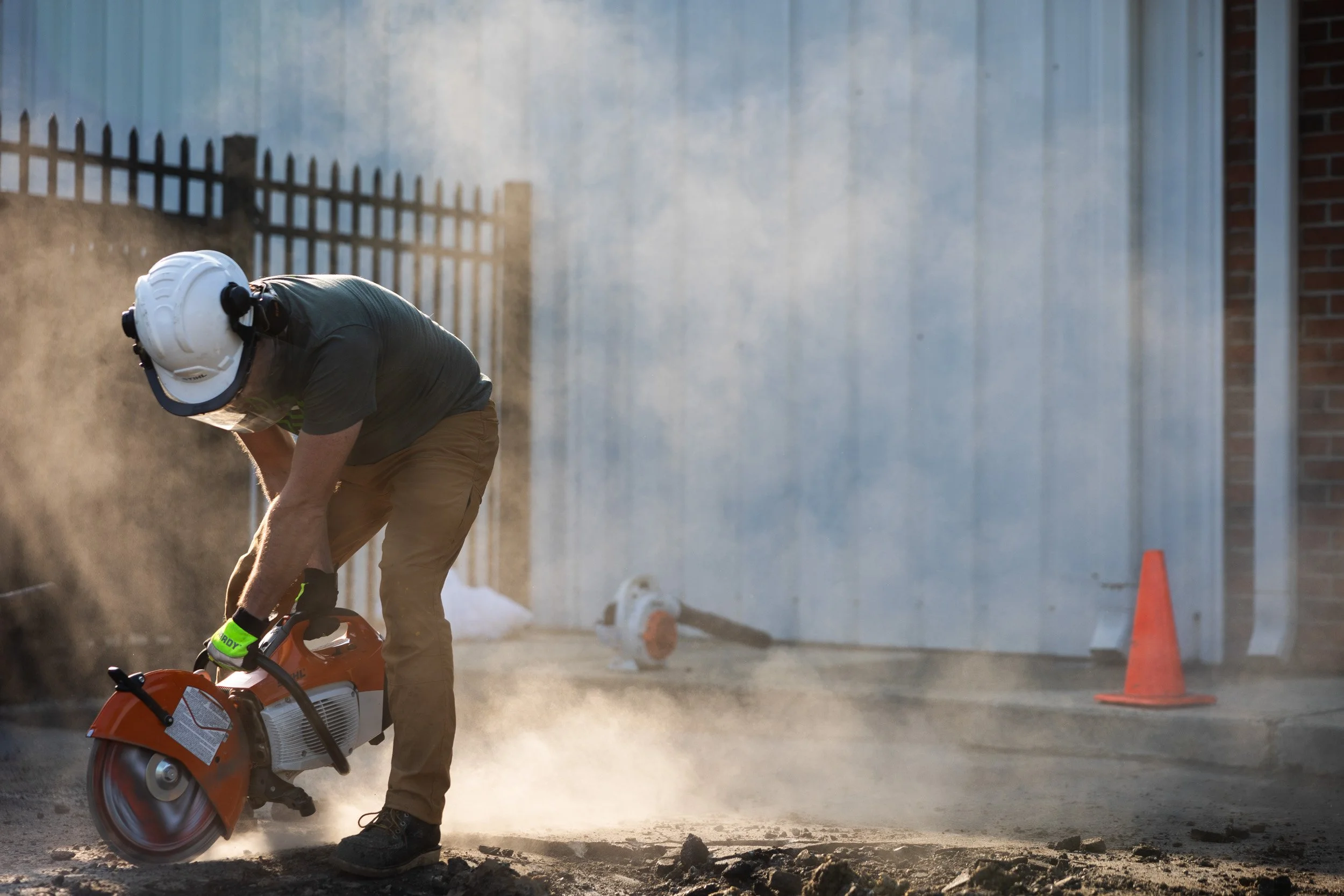 A worker wearing safety gear, including a helmet and gloves, is operating an orange power saw on an asphalt repair site, cutting through an asphalt surface with dust and debris in the air. Orange traffic cones are visible in the background.