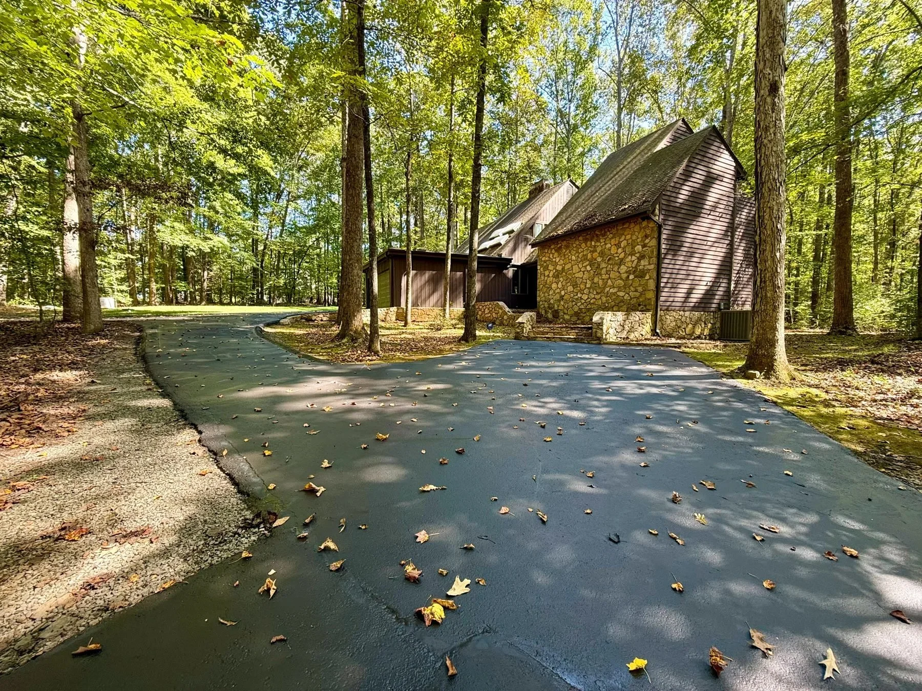 A freshly sealcoated driveway curving through wooded grounds toward a stone-and-shake building, with fallen leaves scattered across the dark pavement.